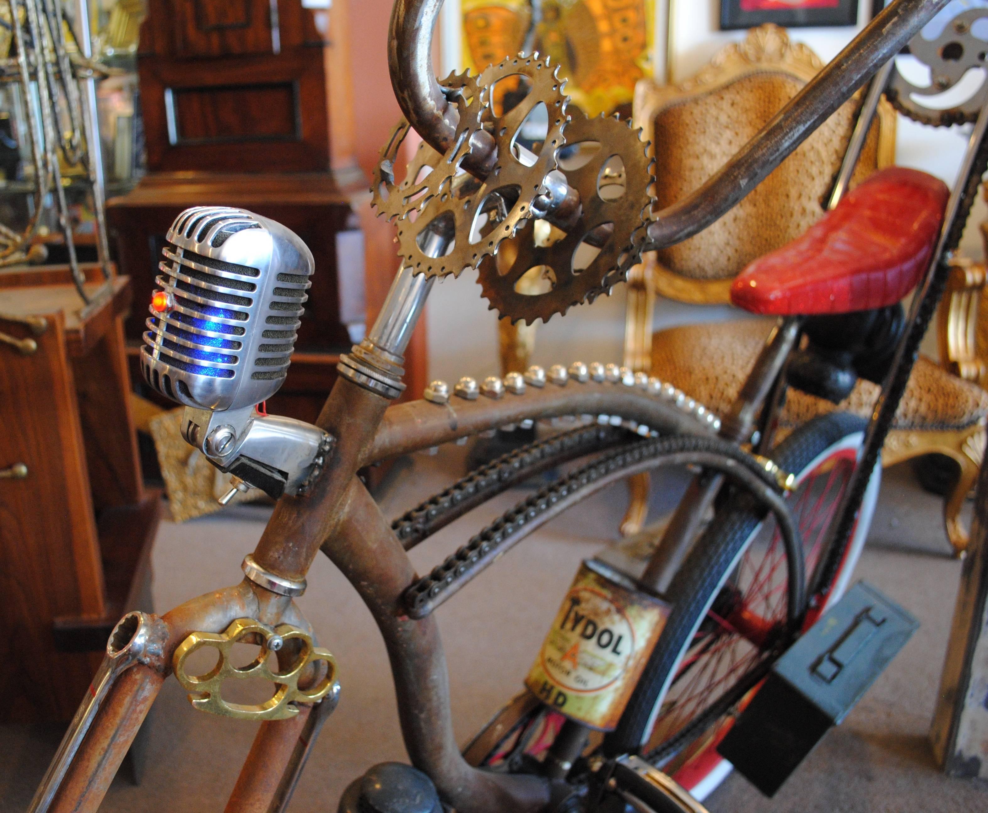 This custom built bicycle has so many random features that it takes on the cohesive design of a rat rod. Some of these features include: Brass knuckles, ammo box, welded chain, cross mirror, lanterns, oil can and a vintage microphone. The microphone