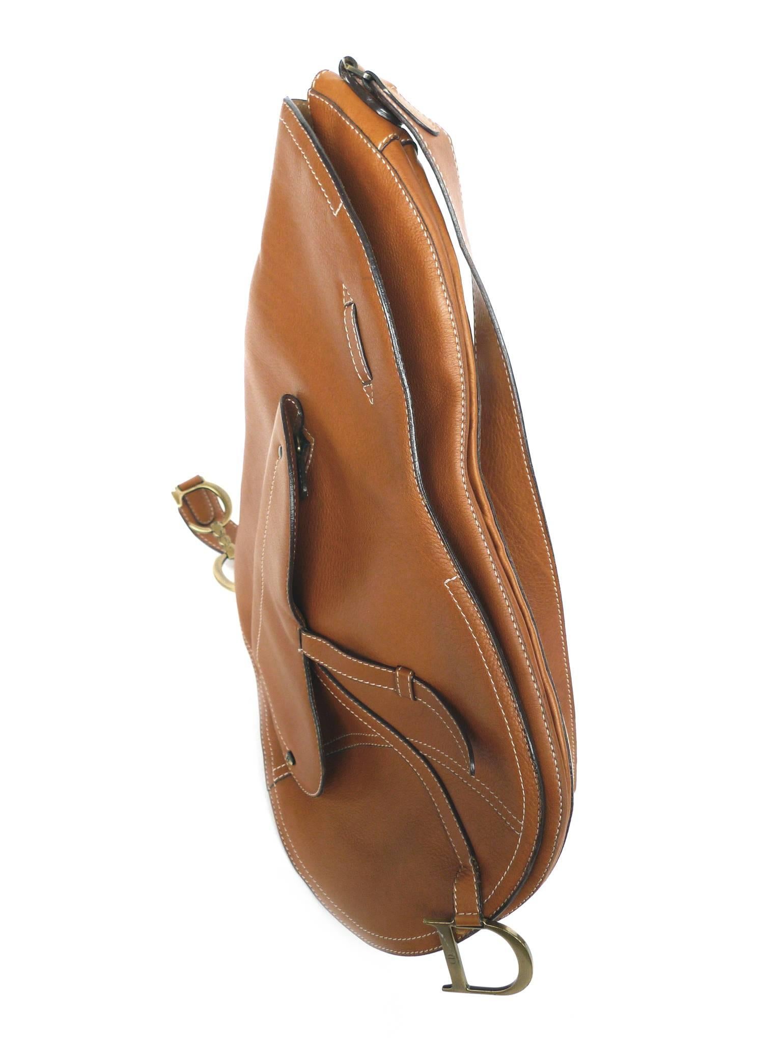 This saddle bag by Christian Dior is crafted from tan leather. Its exterior features include contrast-stitching in a canvas hue, gold-toned brass hardware, and a flat shoulder strap. The interior flap is lined with suede while the interior itself is
