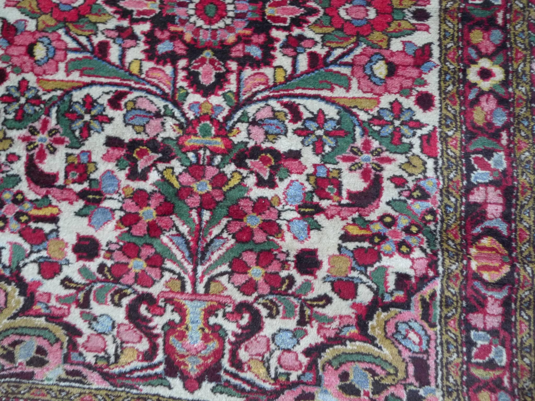 This Persian rug is small and woven with thick wool. Its design consists of central floral motifs in a white field. A dense palette of green, pink, brown, black, and white hues is repeated throughout and in the border pattern.