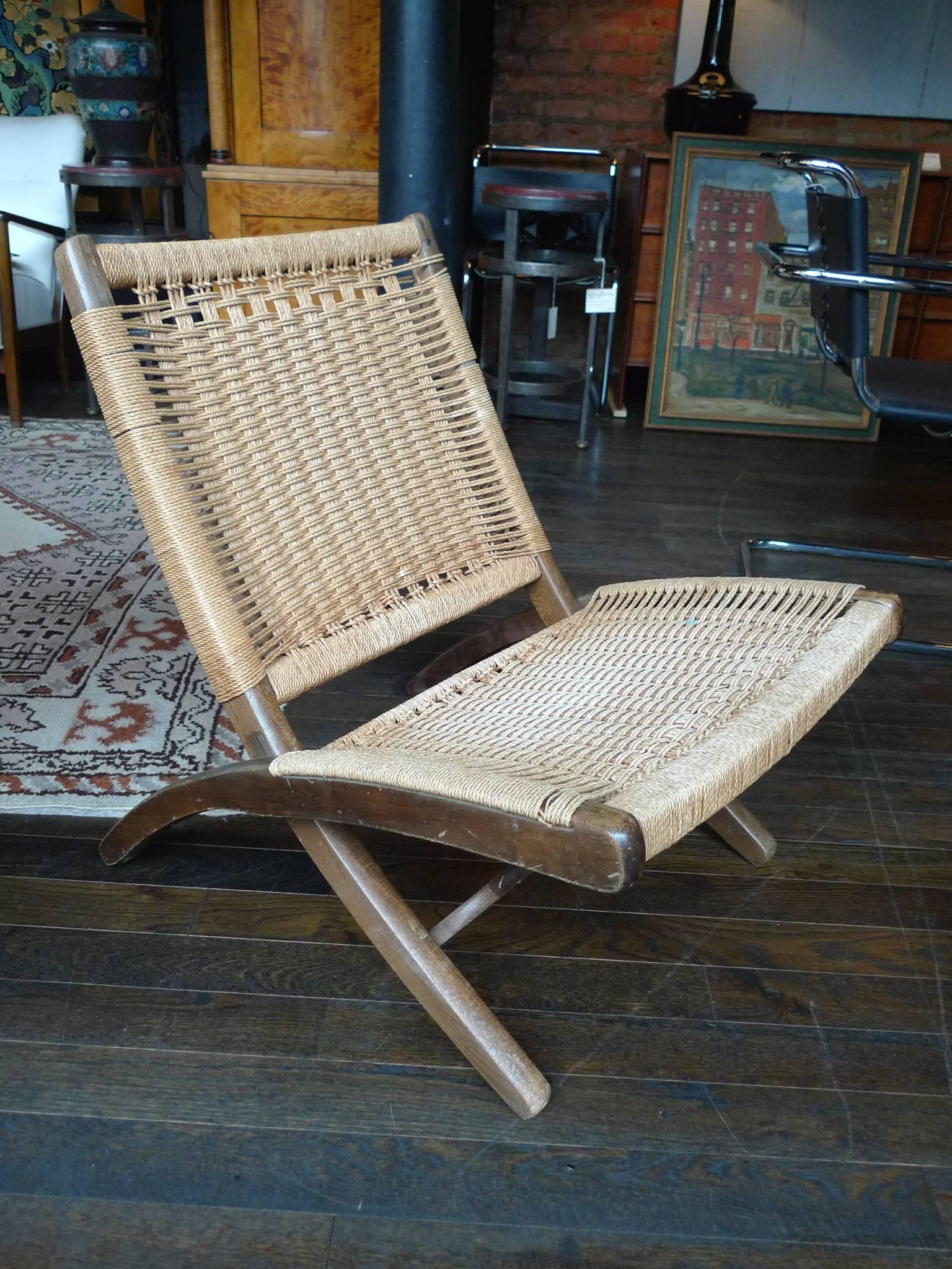 This Mid-Century Modern folding chair is in the style of Hans Wegner. Its dynamic design consists of a crisscrossing diagonal walnut frame. Sturdy rope is woven across the frame, creating the back and seat. The walnut has beautiful rounded edges.