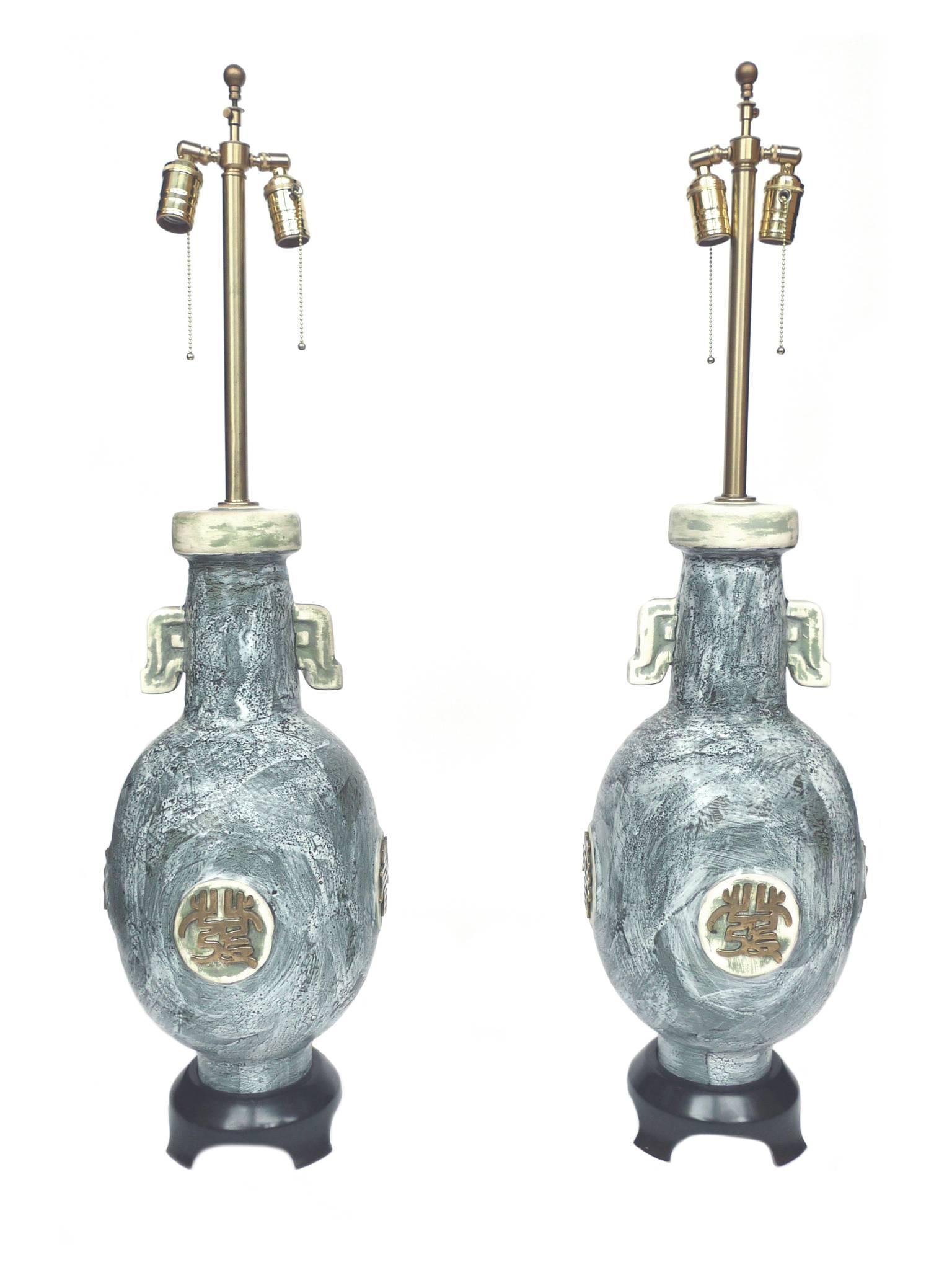 These lamps were produced by the Marbro Lamp Company. Their design includes a ceramic jar body with a painterly blue-gray glaze and stylized brass decorations adorning each side of the lamp. The bases are ebonized wood. The lamps have been newly