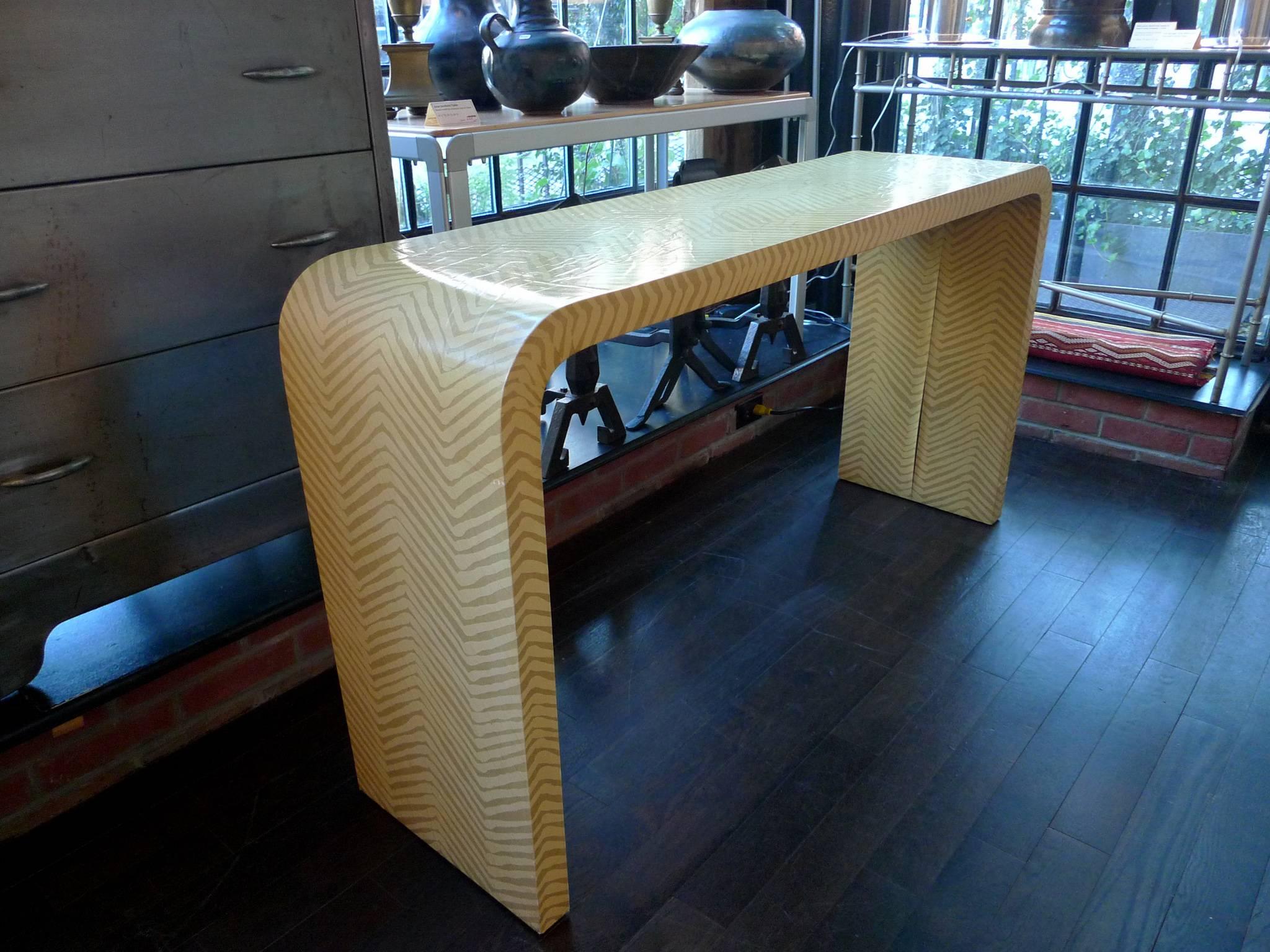 Similar to the designs of Karl Springer, this console table combines a Minimalist shape with visually rich texture. The curved frame measures approximately two inches and is covered in a grasscloth-like material set in yellow stripes. It's a simple