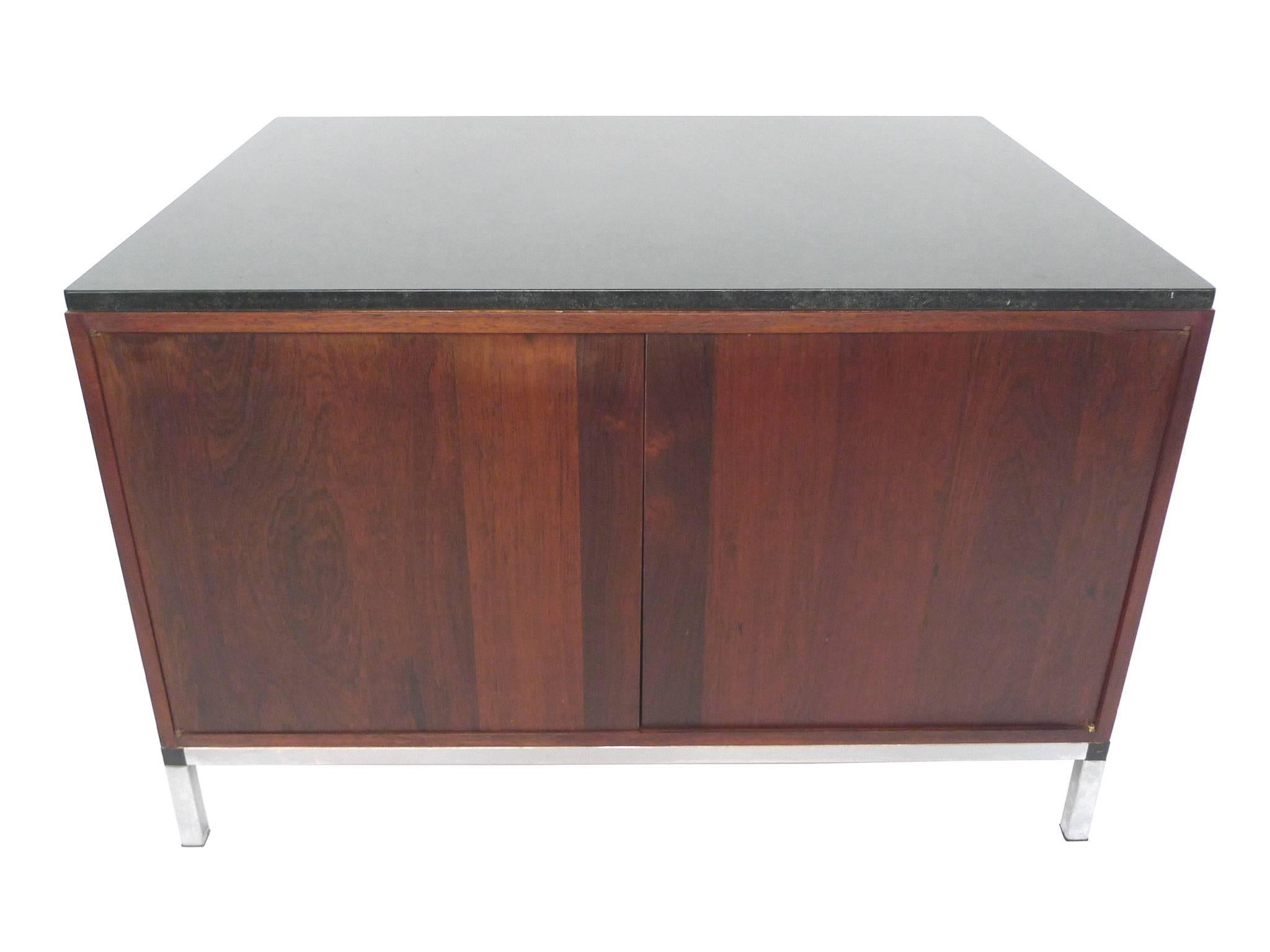 This Midcentury cabinet is in the style of Florence Knoll, whose Minimalist designs made use of simple shapes and clean lines. The cabinet consists of a black marble top, a rosewood body, a chrome base, and brass hardware. The rosewood is a rich