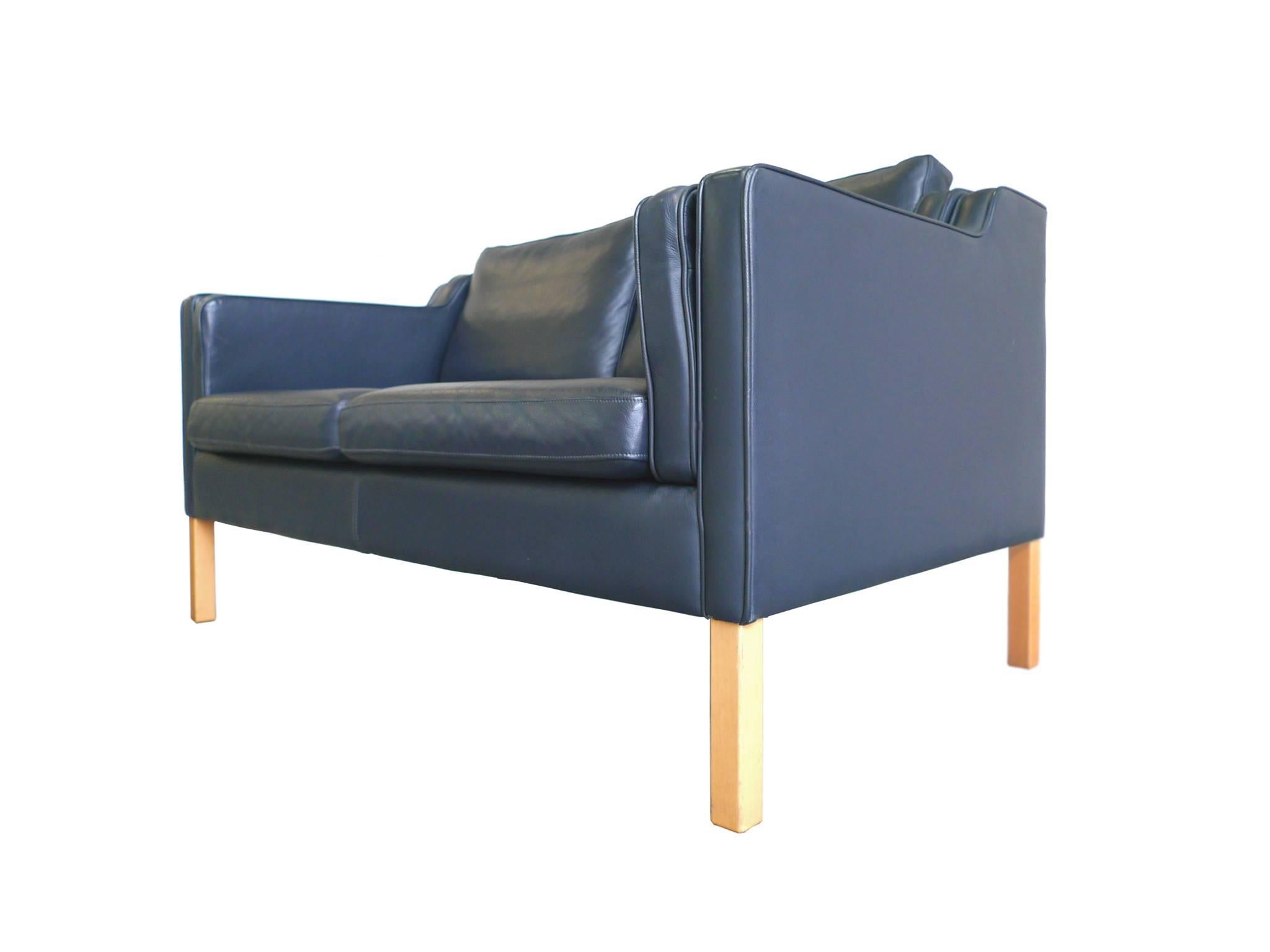 This settee is in the style of Børge Mogensen and was manufactured by Stouby. Its design is one of clean lines and soft, rounded edges. The upholstery is a dark cobalt blue leather, while the frame is beech wood. The settee has a low back and