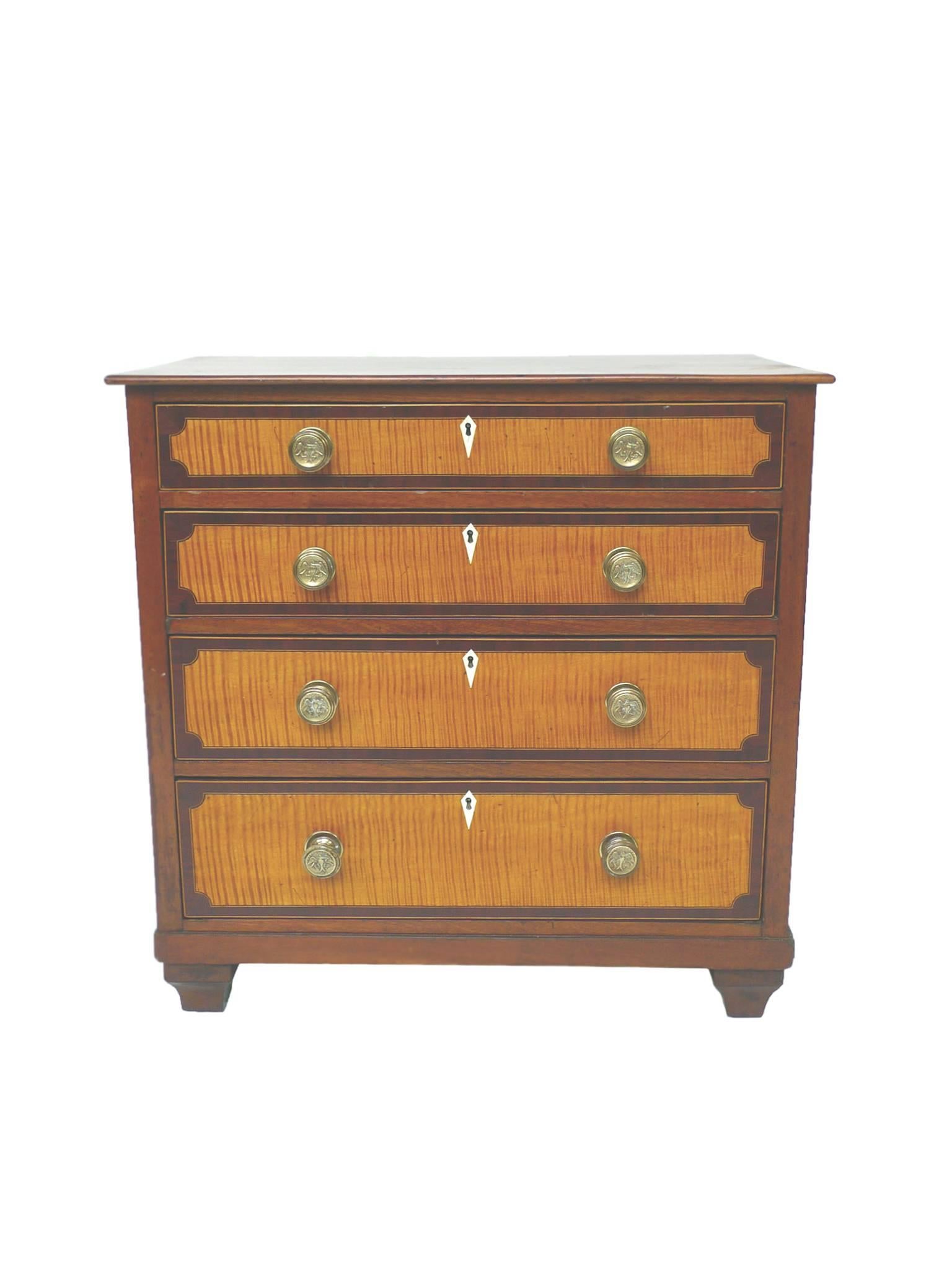 This Federal-era case piece is crafted from maple wood. The design includes four graduated drawers with flame tiger maple fronts, round pulls with an embossed bird emblem, and square turned bun feet. This modest-sized bureau has been refinished,
