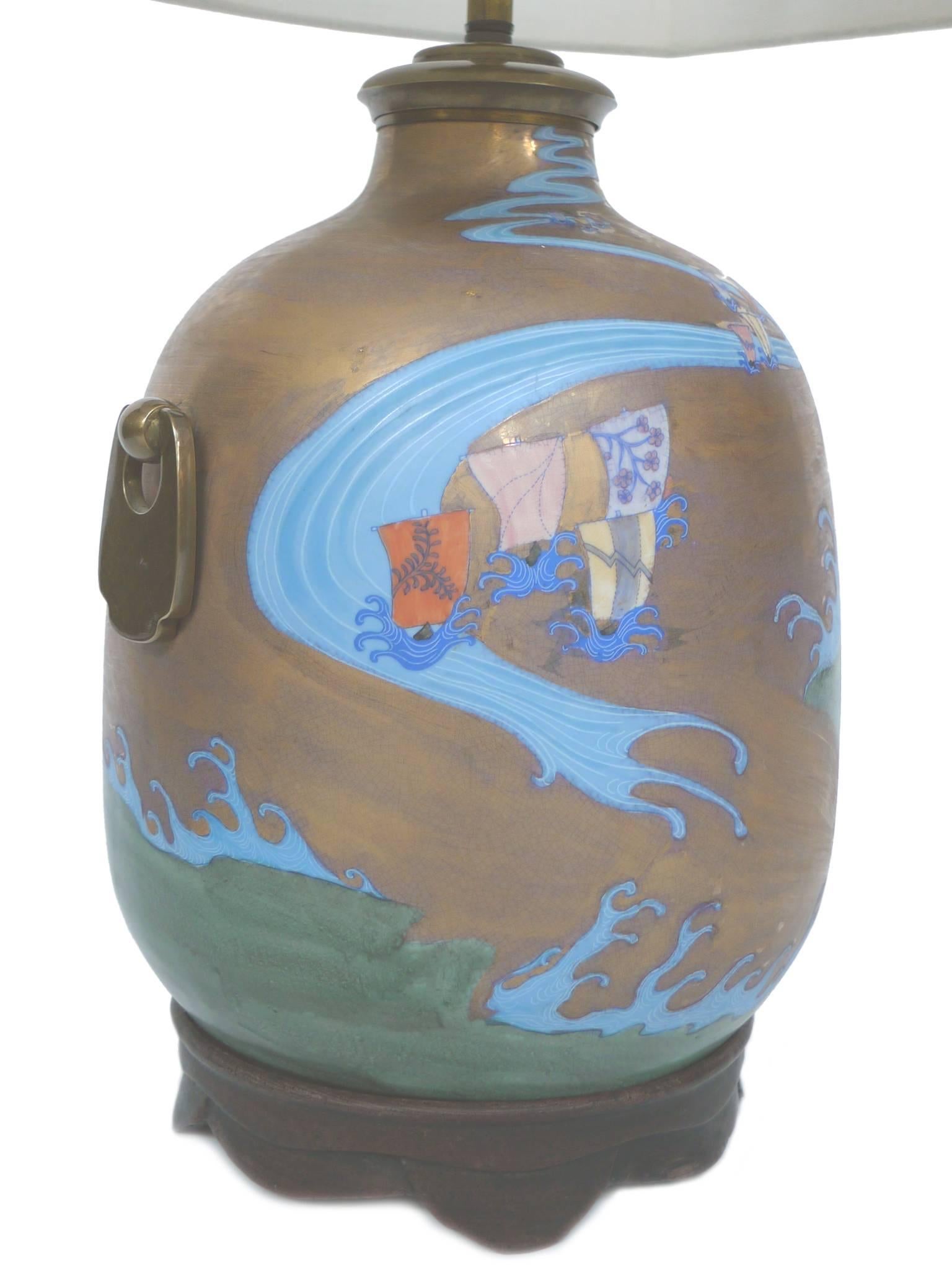 This 20th century table lamp has an oblong brass body with a wood base. The brass body is decorated with a wispy, stylized maritime scene, painted in enamel and wraps around the lamp. There's a brass handle at each side. The wood base is carved with