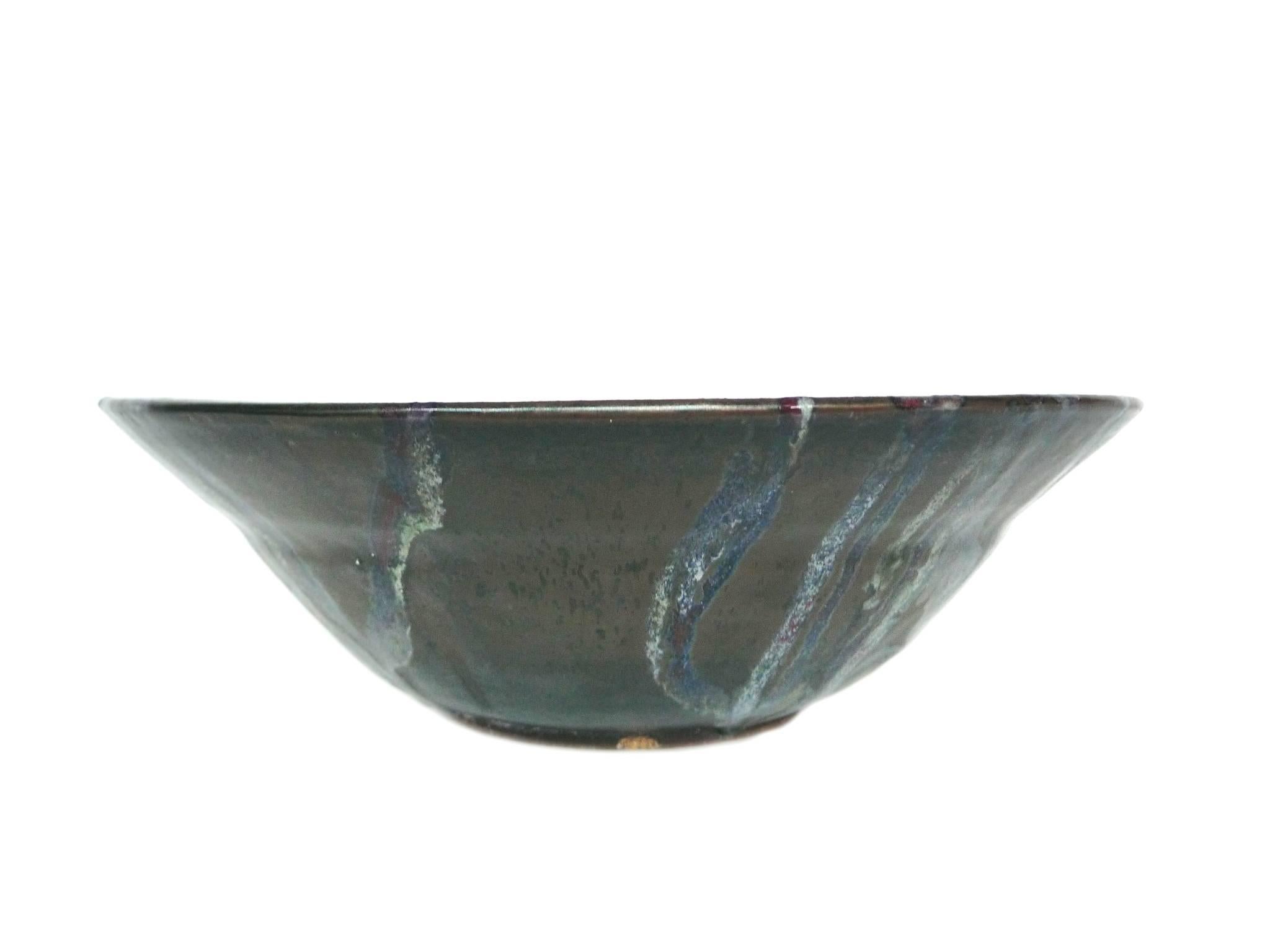 This Thom Lussier bowl comes from a series of ceramics that combine a variety of textures and glazes, evoking geological forms. The surface is painted and layered with green and stone-grey hues, with hints of blue. The centre is glazed with alizarin