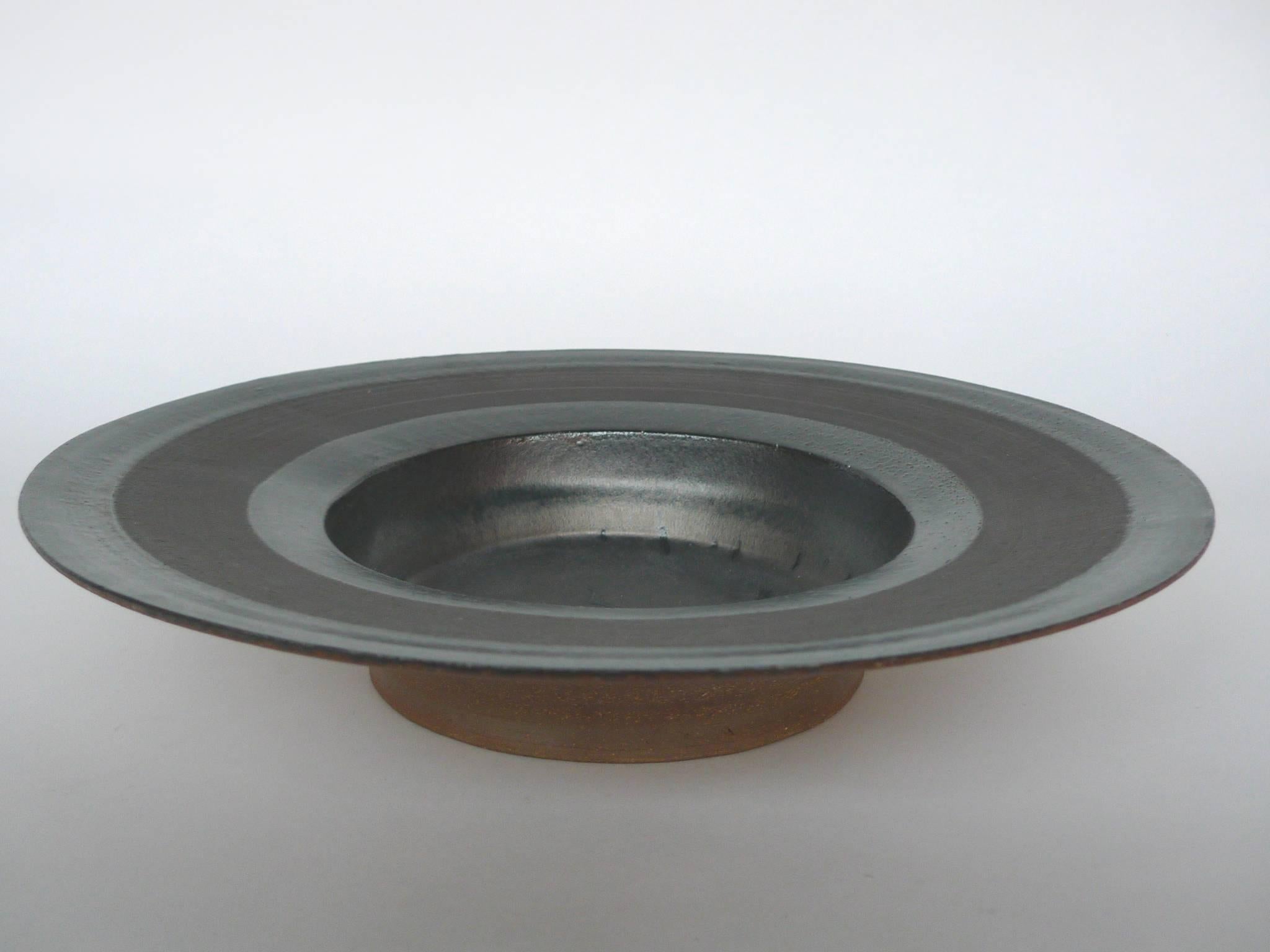 This ceramic saucer bowl is by Thom Lussier. It's part of a series of pottery on which the artist applies a mixture of glazing techniques to create richly textured surfaces. The top and interior sections of this saucer bowl combine metallic and