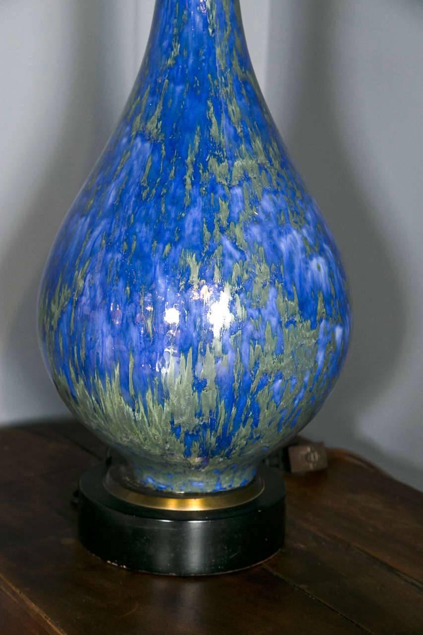 This 1960s Italian lamp is ceramic with a scintillating blue and gray-green glaze. It's a tear drop shape with a long stem. The top portion is brass, while the base is black-painted wood with brass trimming. The lamp is rewired and refurbished with