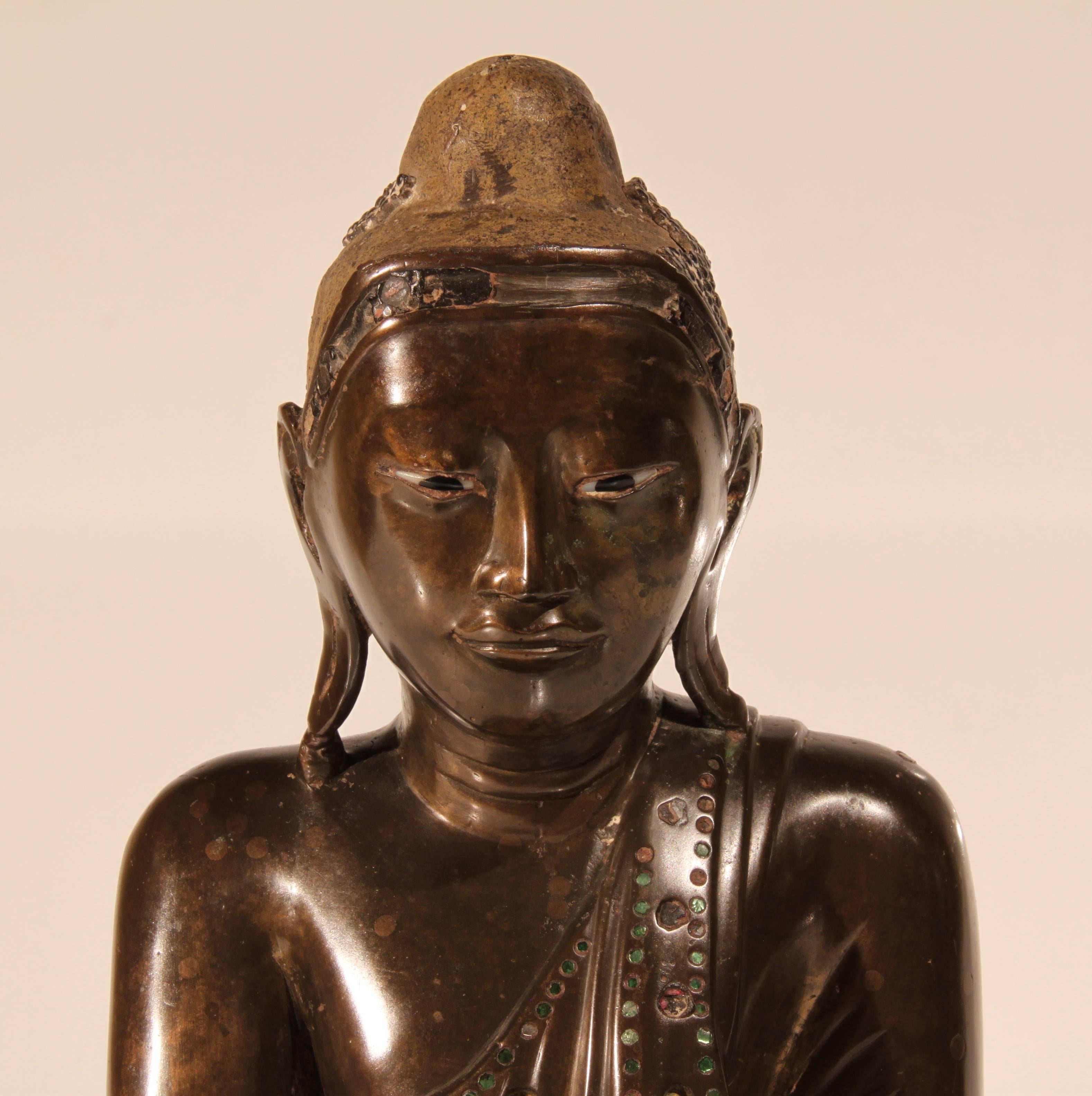 A mid-19th century Mandalay period Thai bronze Buddha inlaid with green glass and having enamel inlaid eyes. The head dress once lacquered and gilded, now with traces of lacquer. The left hand cast separately and held in place with lead. Many