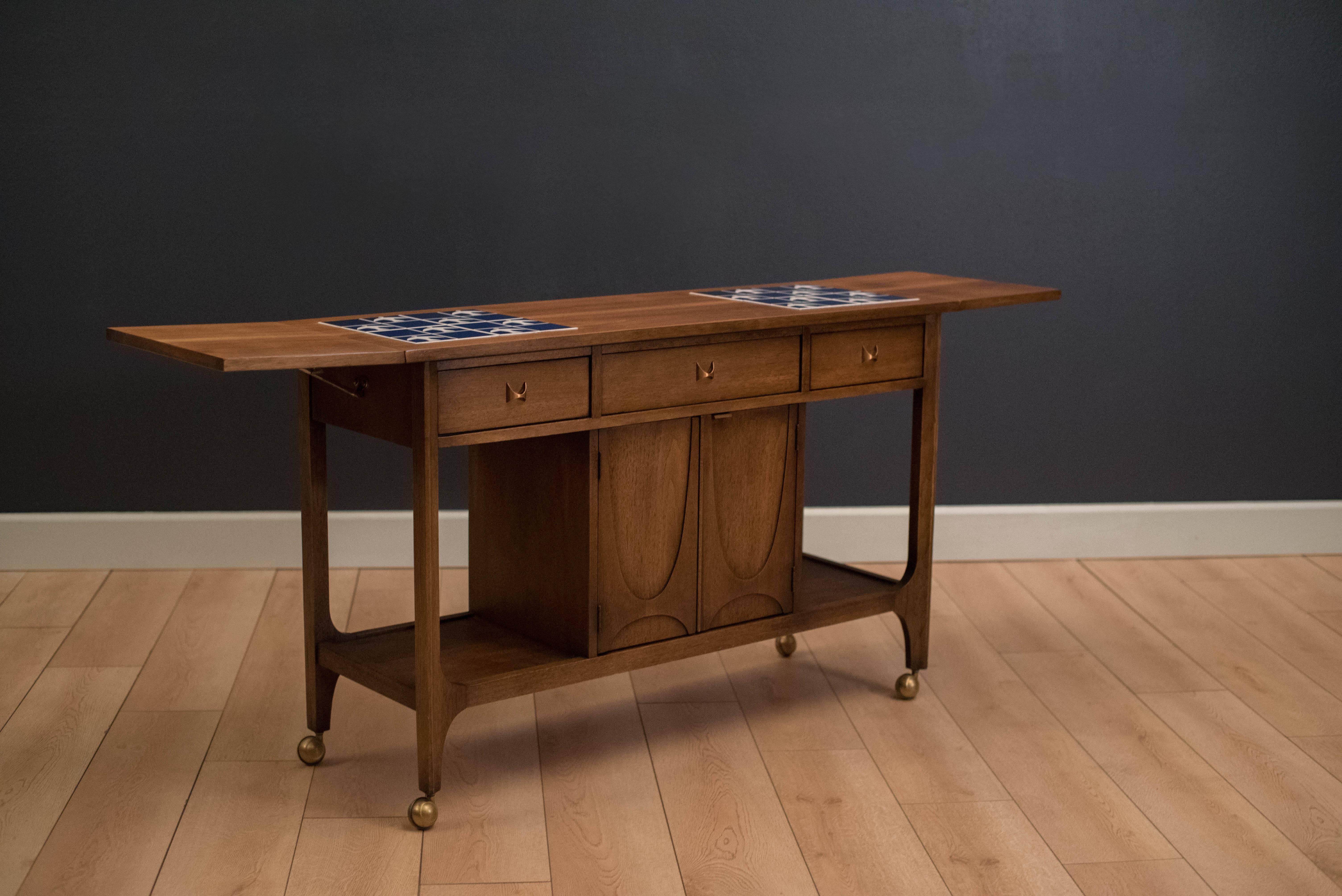 Vintage Brasilia bar cart manufactured by Broyhill Furniture Co. in walnut. This piece includes three drawers and a middle cabinet offering plenty of storage. Drop leaves extend to increase the work surface area and features the line's signature