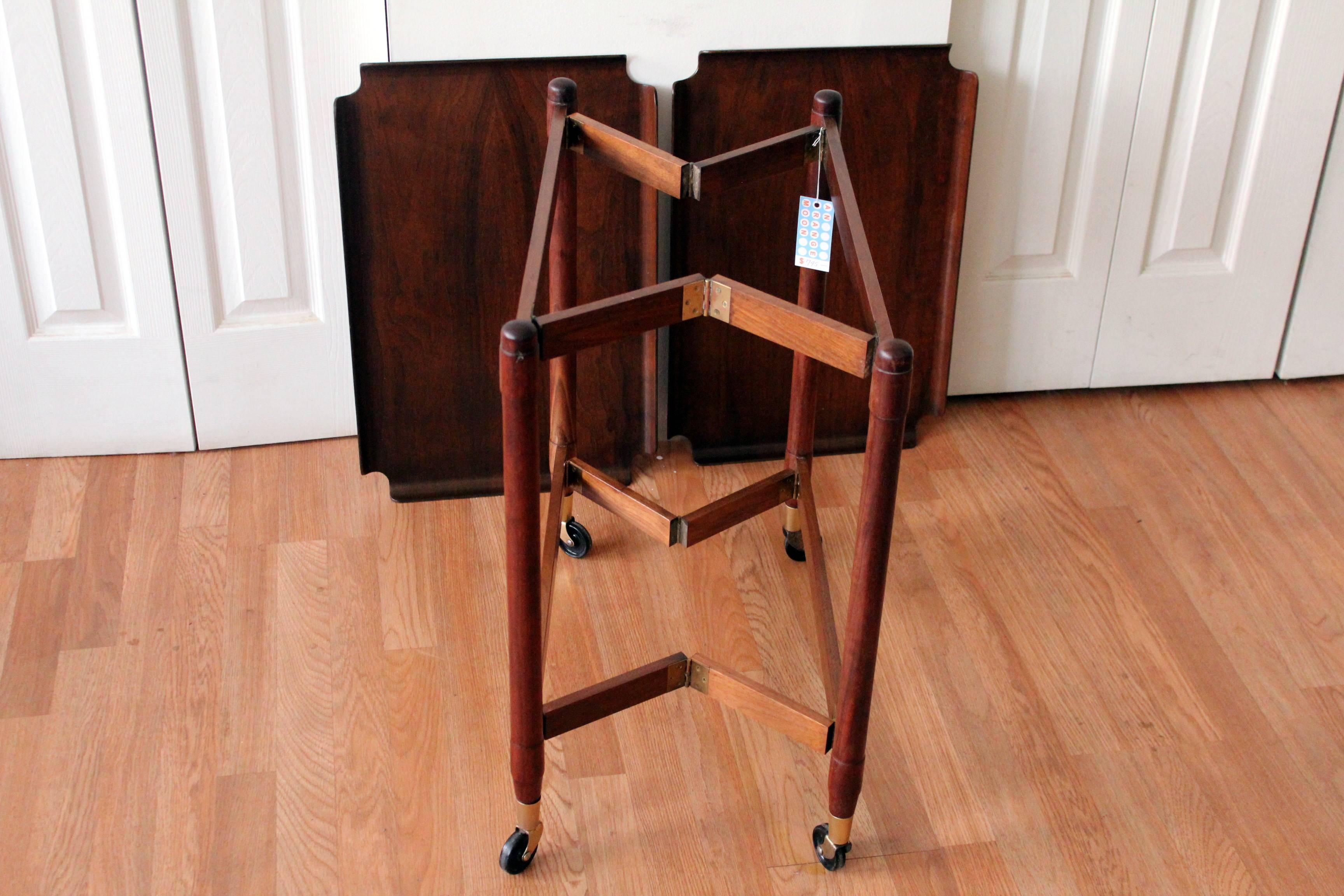 This well designed Mid-Century Modern John Stuart trolley bar cart in walnut is perfect for entertaining. Notice how beautifully it folds for storage? The cart sits atop wheels with brass caps and hinges. It also boasts removable trays perfect for