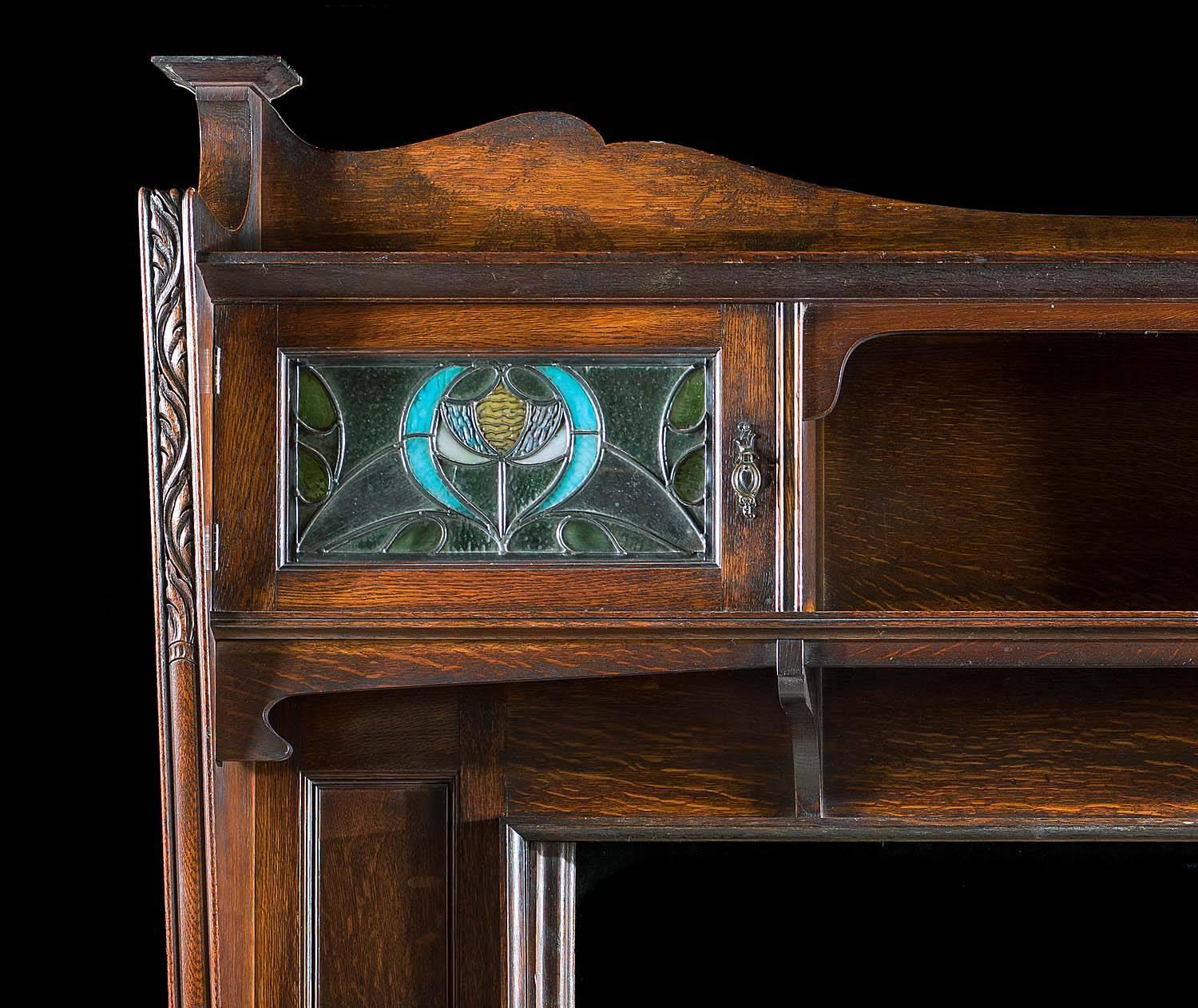 An Arts & Crafts oak fireplace surround in the manner of the Cheshire designer
George Faulkner Armitage (1849-1937), with a deep moulded shelf above a pair of cupboards with stained and leaded glass doors set either side of the open lower shelf