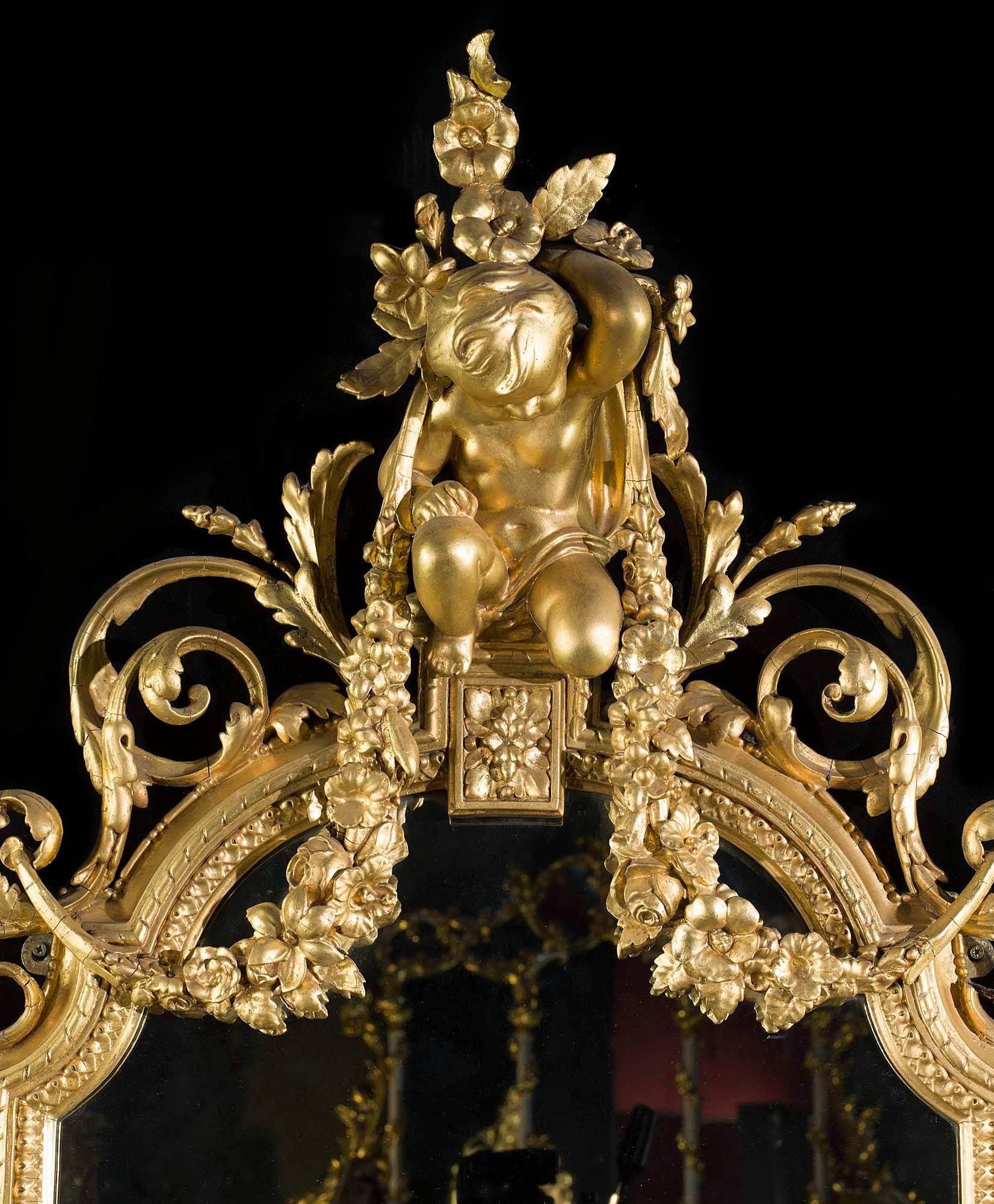 A sumptuous antique giltwood and gesso Louis XV style girandole mirror. The generous pediment is in the form a single putti holding aloft garlands of summer flowers among scrolling foliage. This is set above the arched topped mirror with its