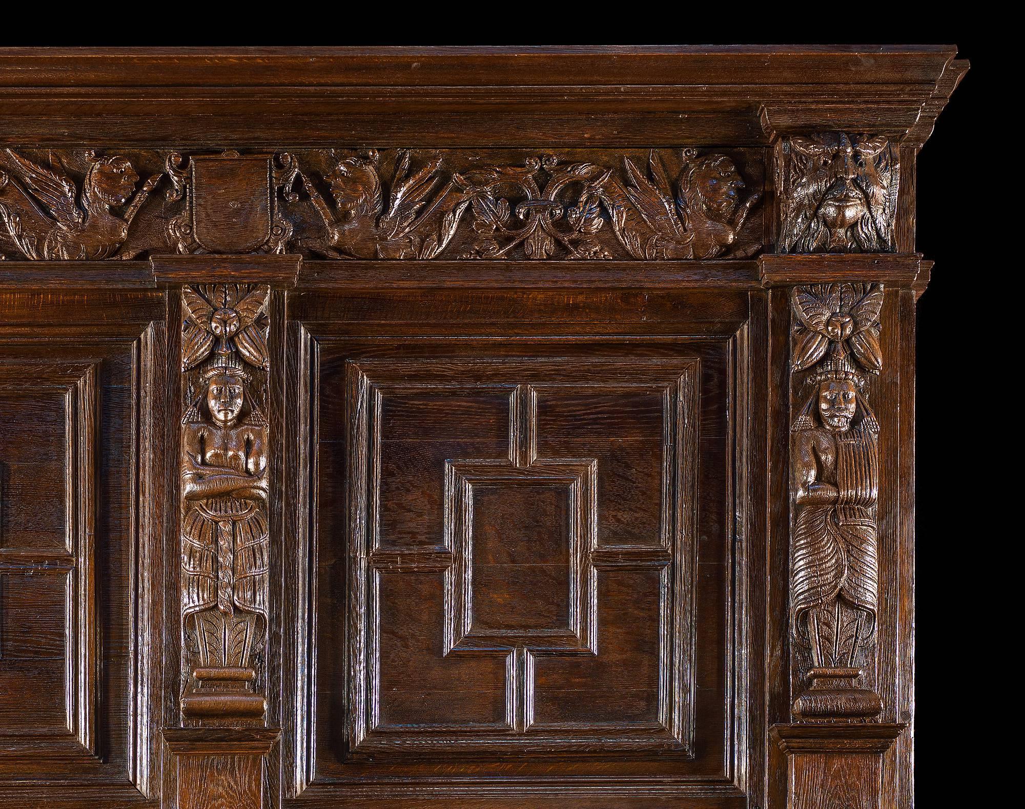 A tall carved oak antique fireplace surround and overmantel. The surround with fluted Corinthian column jambs and a cushioned mantel shelf, the overmantel with two quartered panels separated by three pilaster Aztec style figures with stylized floral