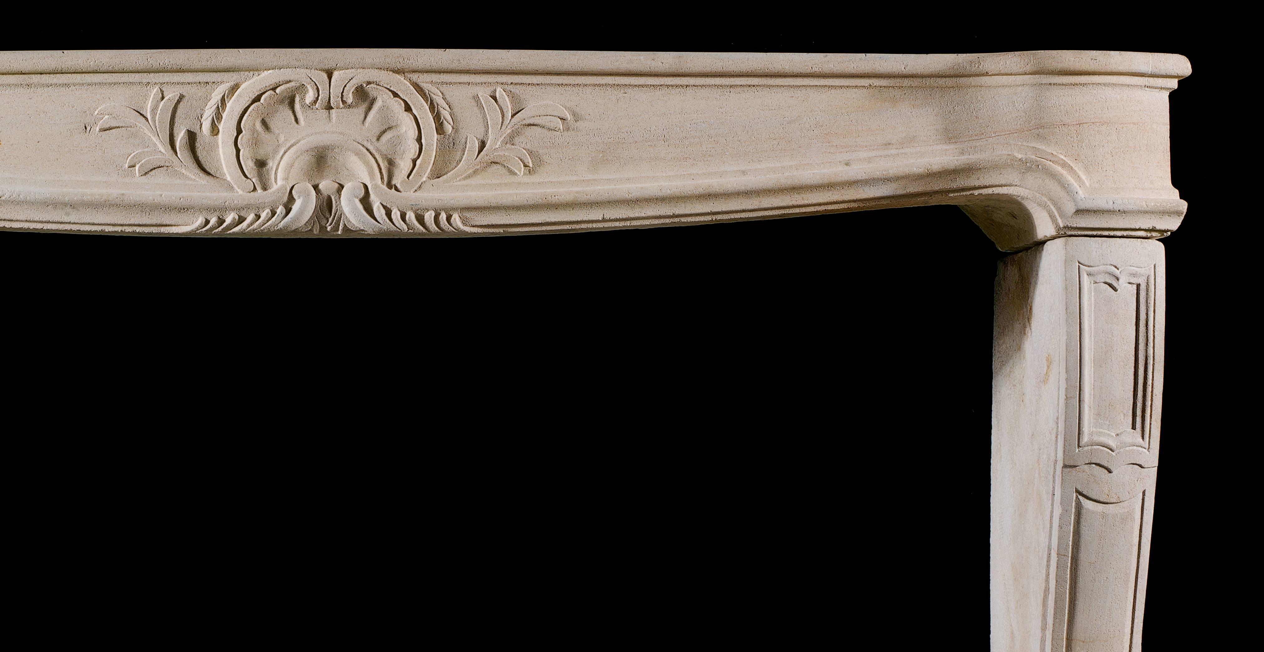A French Rococo antique fireplace mantel in the Louis XV manner, carved in limestone. The serpentine shelf is integral with the similarly formed frieze, carved from the solid and decorated with a simple floral shell cartouche, supported on panelled