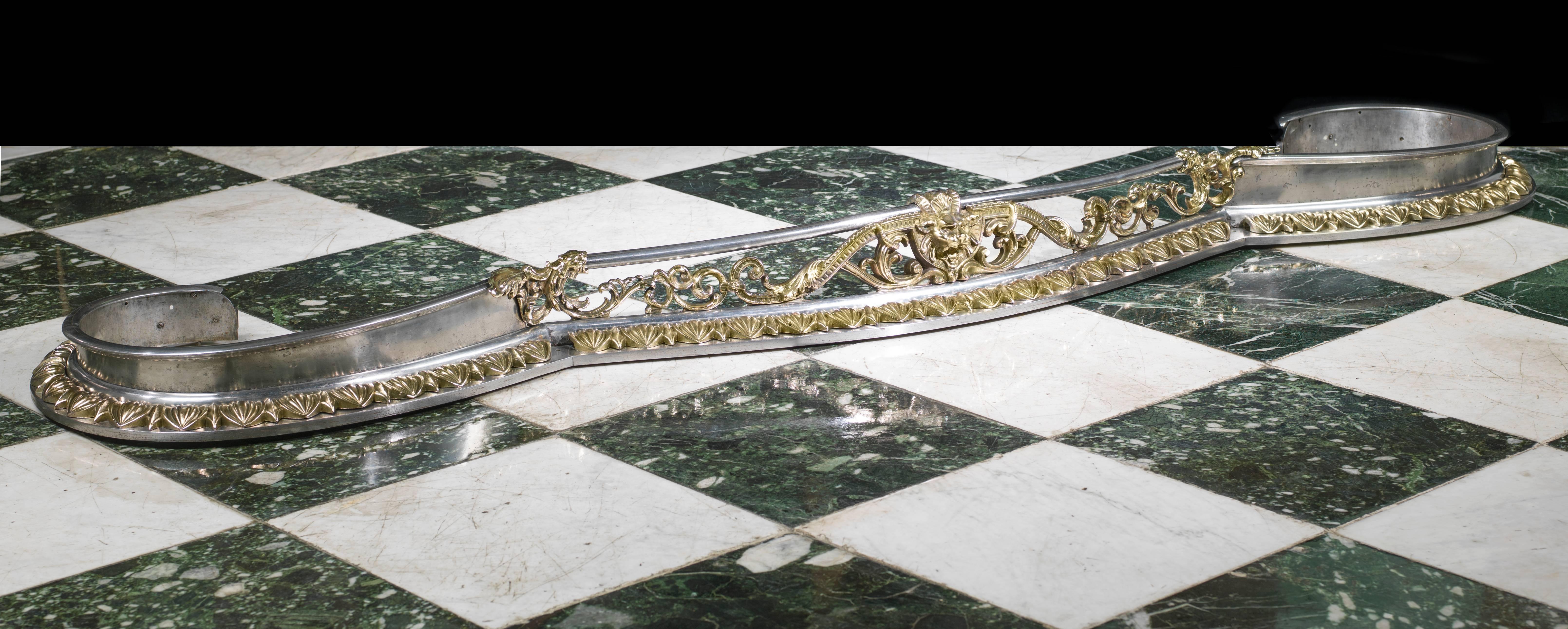 A substantial cast iron and brass mounted Victorian serpentine fireplace fender, with continuous cast brass detail along the base and a horned grotesque mask centred on the bar held either end in the mouths of mythical creatures. 

English, circa