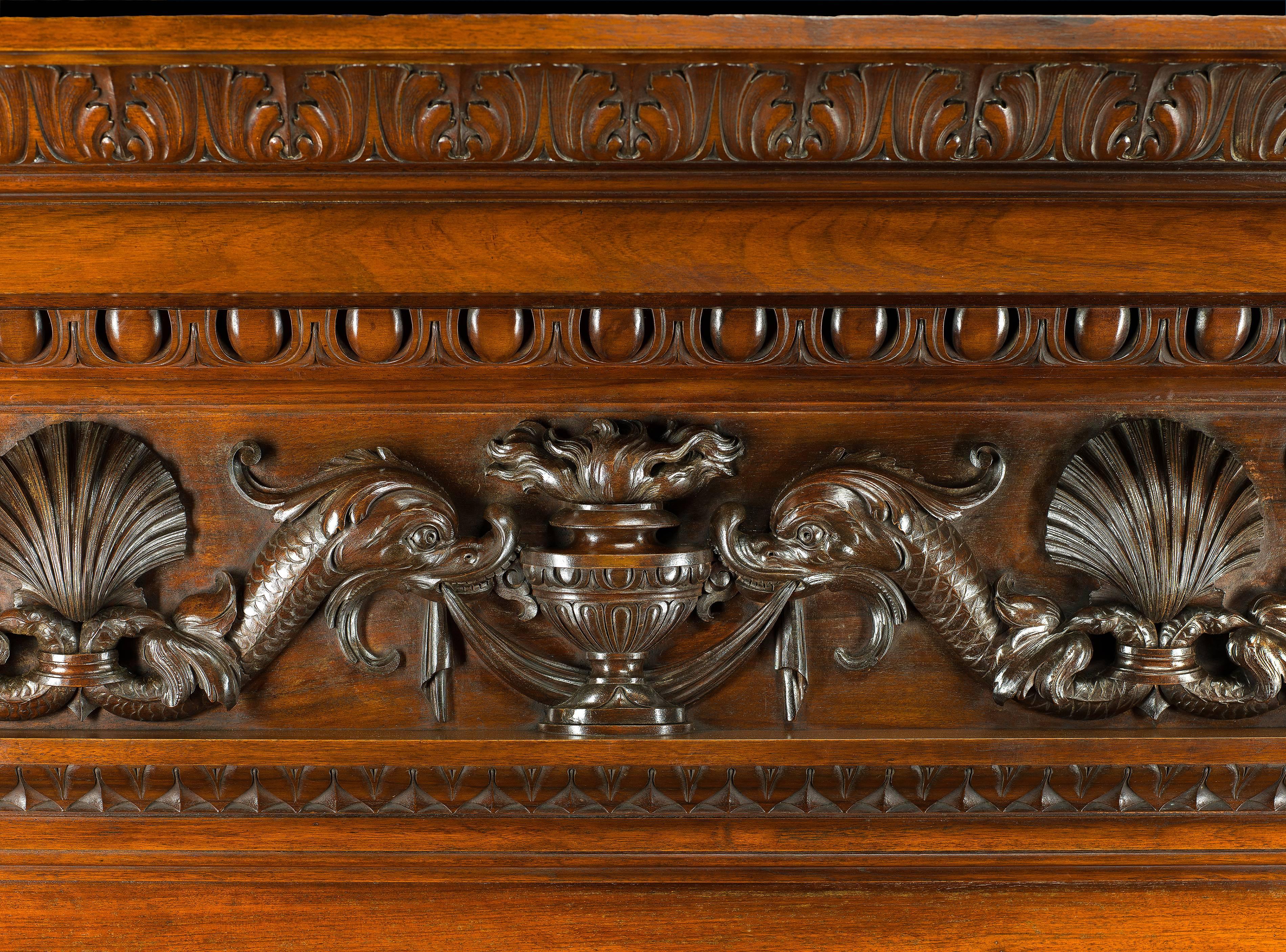 An enormous and grand antique walnut chimneypiece in the neoclassical style. The frieze elaborately carved with three pairs of sea serpents alternating with flambeau urns and scallop shells. The shells are repeated on the panelled surround of the