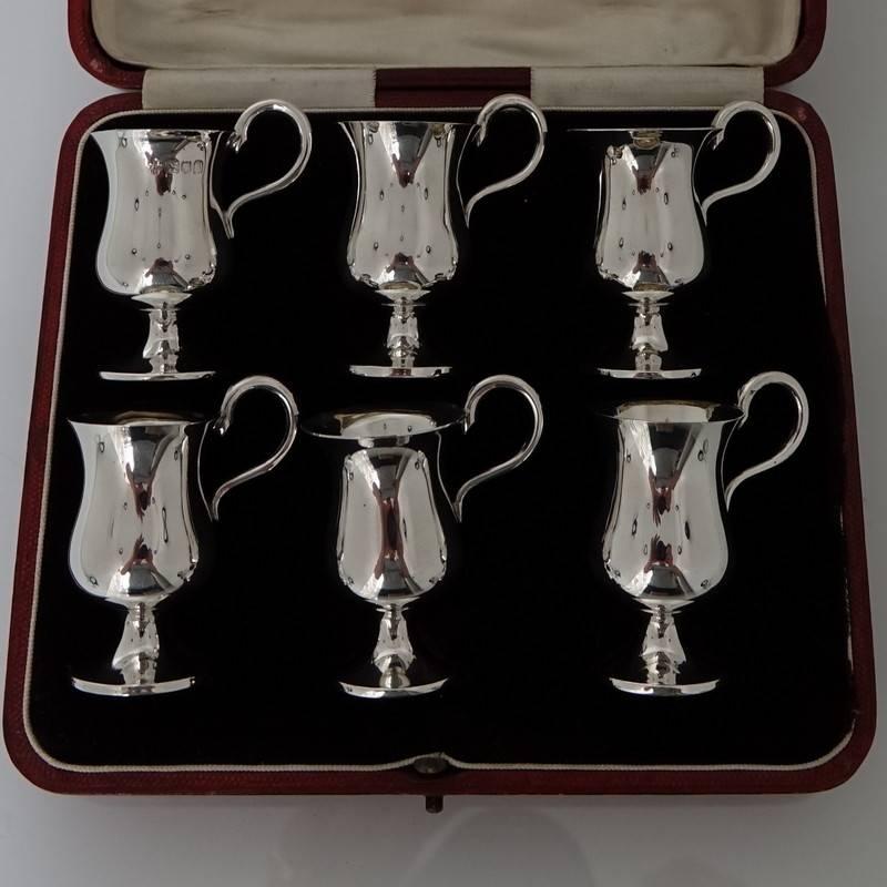 A delightful set of six plain-formed tot cups styled in the manor of a traditional tapered in bellied pint mug. The handles single scroll and the body sit on a circular raised pedestal foot. There is a velvet lined red presentation box.