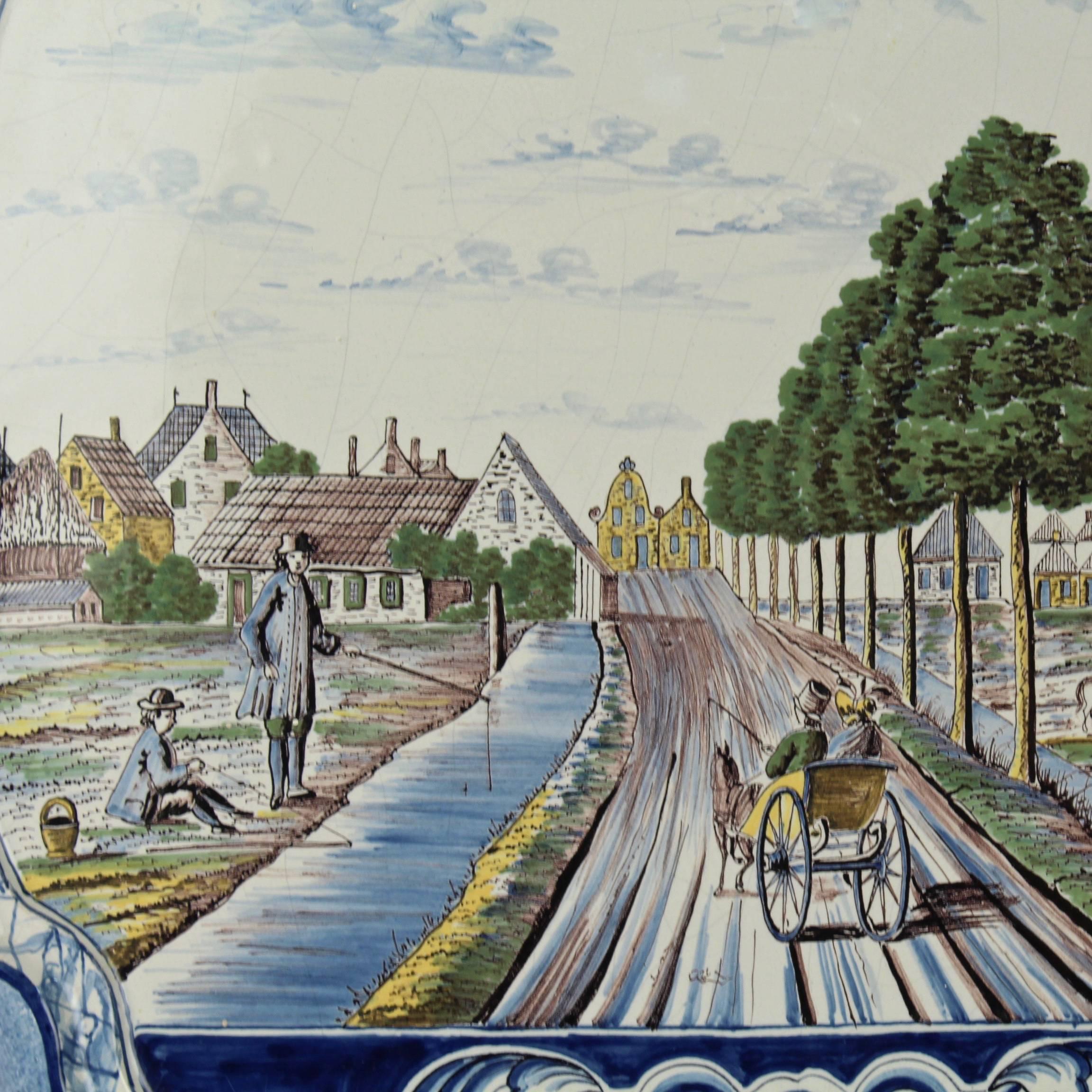 A large polychrome Dutch delft pottery wall plaque. 

It depicts a scene with a carriage passing through a bucolic landscape on a road flanked by a canal and pastures en route to a town. 

Dates to late 19th or early 20th century (or possibly