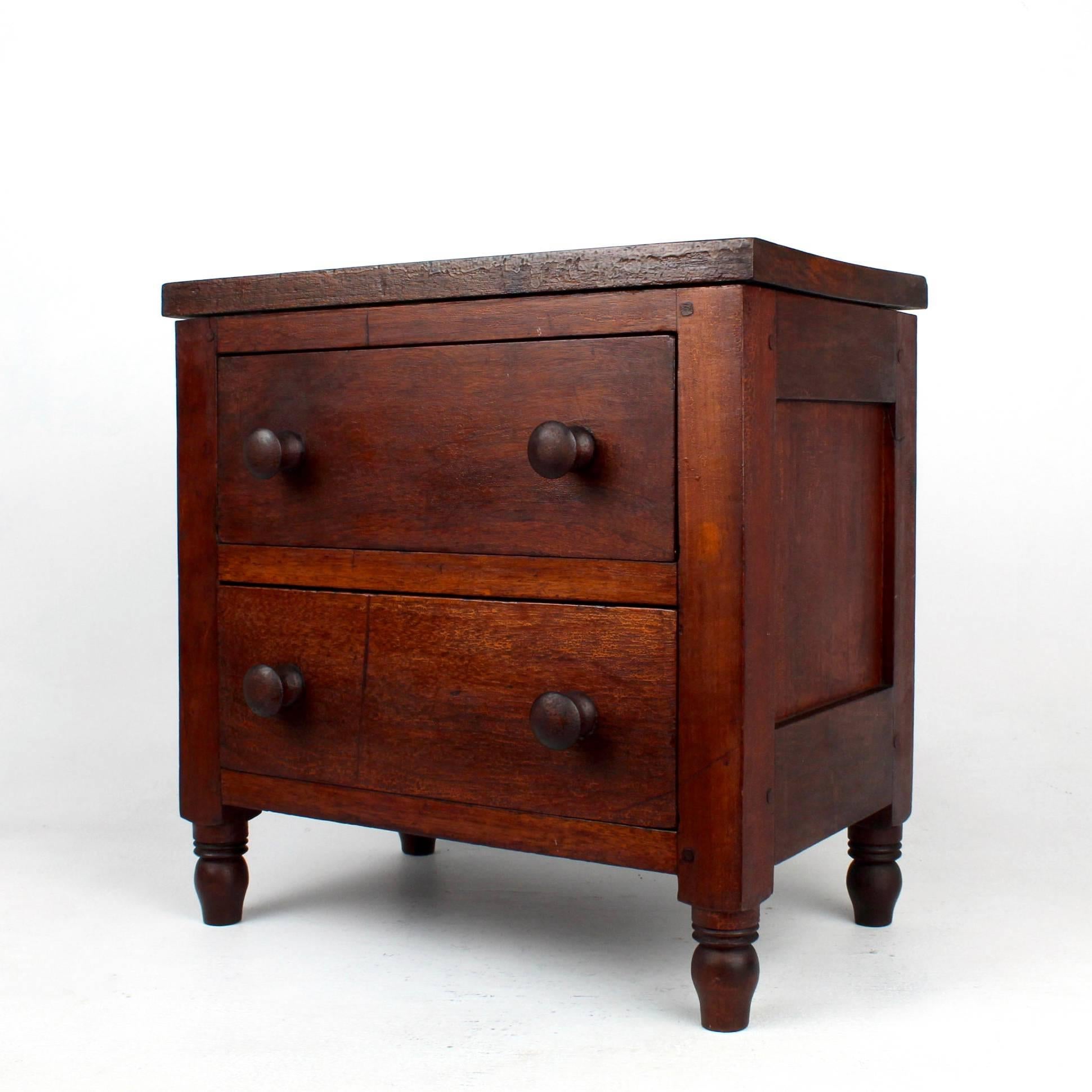 Commode miniature à deux tiroirs de la fin du XIXe siècle.

Les côtés sont recouverts de panneaux et les pieds et boutons sont tournés. 

Composé de noyer, d'une partie secondaire en pin et d'une construction cloutée. 

Trouvé en