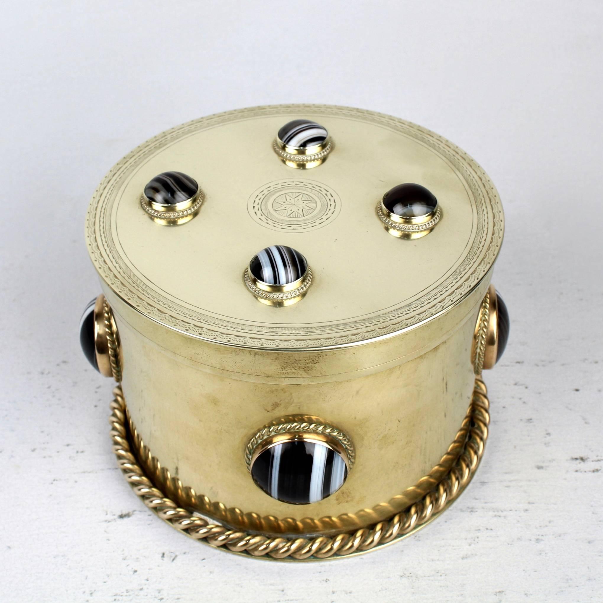 A masculine, round brass box with a wheel engraved lid.

English. High Victorian,

The body and lid are each set with four banded agate (sardonyx) stones cut en cabochon. The slightly extended base supports an applied twisted wire.

Diameter:
