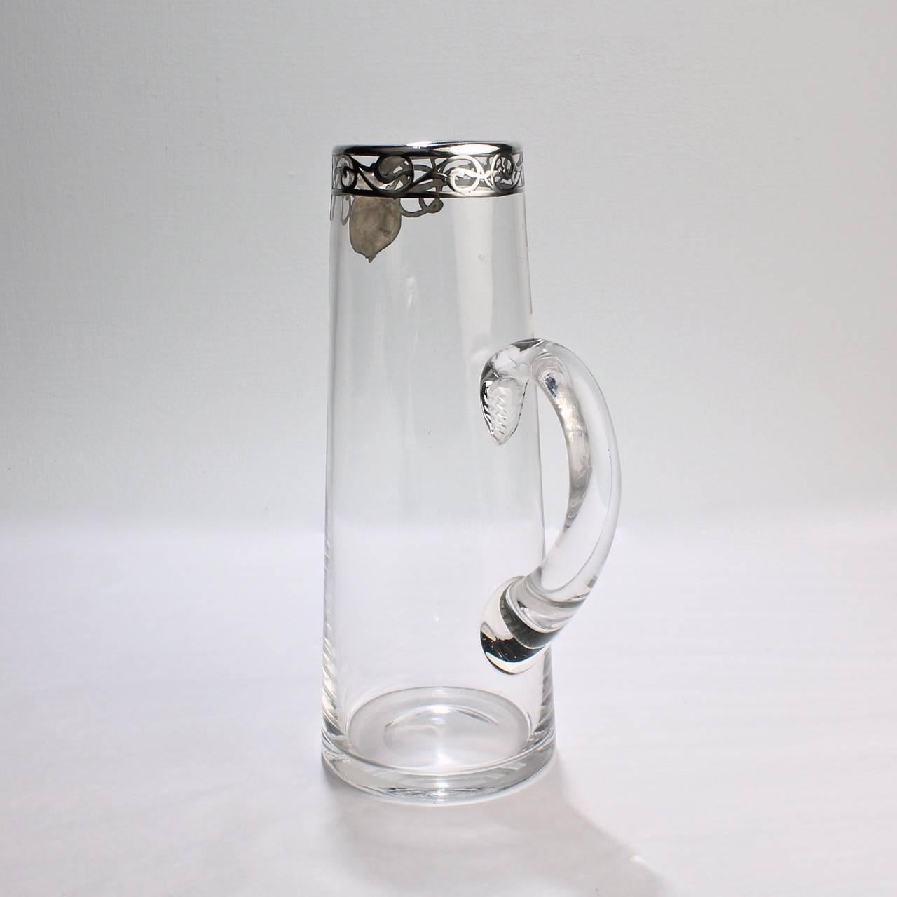 An early 20th century Art Noveau silver overlay glass cocktail pitcher.

With applied C shaped applied handle and silver overlay encircling the top rim.

Base with a polished pontil. Top rim marked Sterling 114.

Height: ca. 10 in.

Items