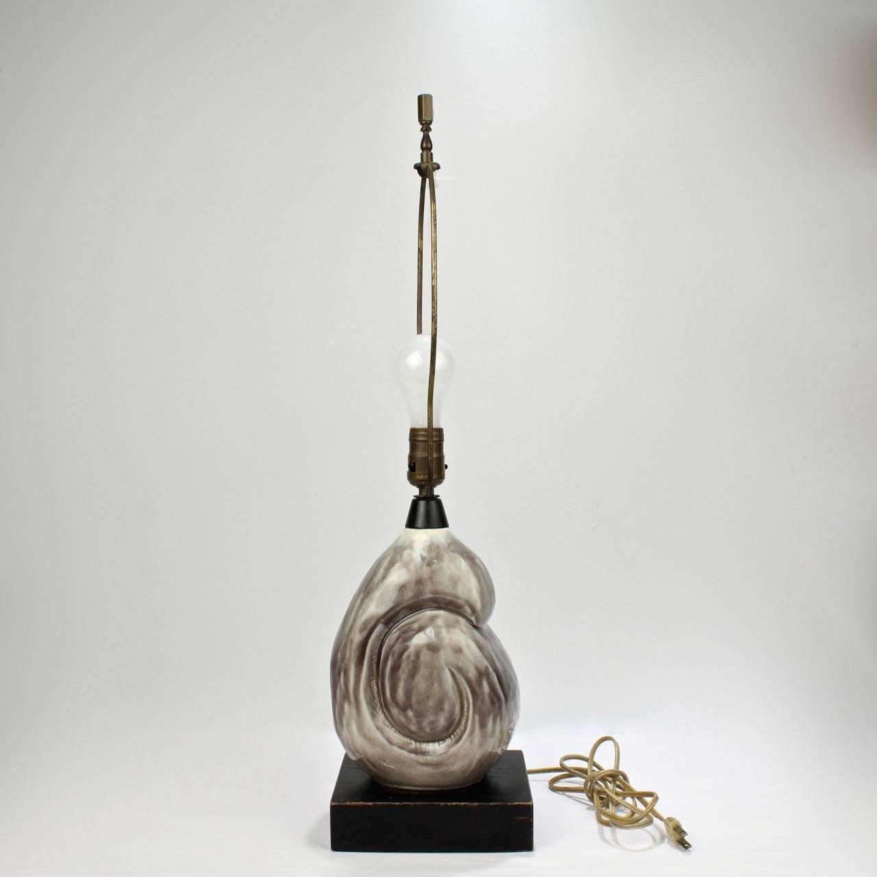 A Mid-Century Modern pottery lamp with a grey-tone nautilus-form top by Yasha Heifetz.

Mounted on a black painted wooden base. 

Stamped Heifetz to base.

Height including fixture: ca. 26 3/4 in.

Retains its original factory wiring and