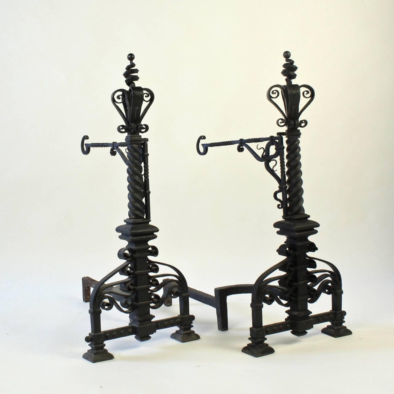 A pair of Arts and Crafts wrought iron andirons with incredibly dramatic scale and weight.

Having a large central twist pedestals supported by ornate scroll-work on square feet, with hinged bracket form arms mounted on the pedestals, and stylized