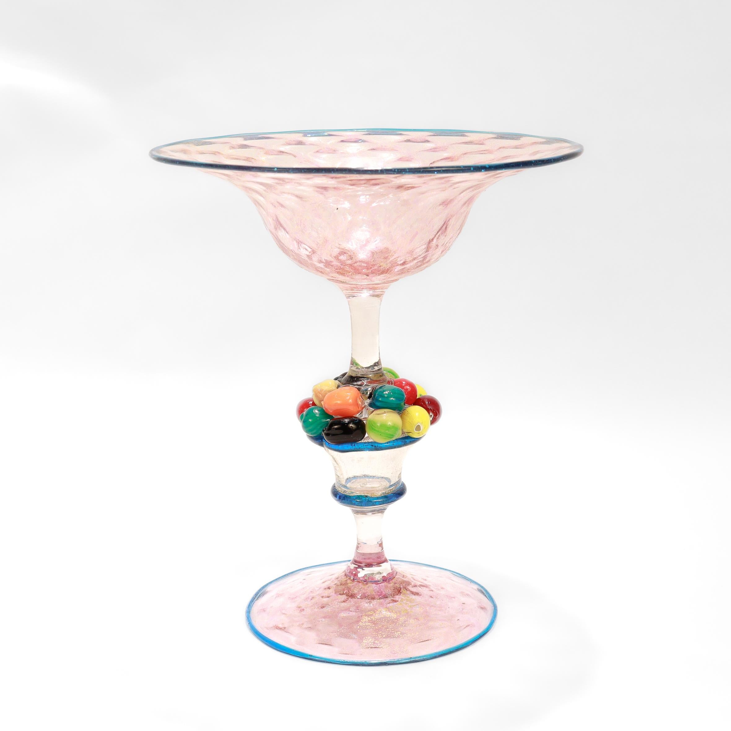 Une compote ou tazza en verre d'art italien.

Attribué à Salviati. 

En verre rose clair avec des accents bleu foncé sur les bords et des inclusions dorées sur l'ensemble.

La tige est centrée sur un panier rempli de fruits miniatures