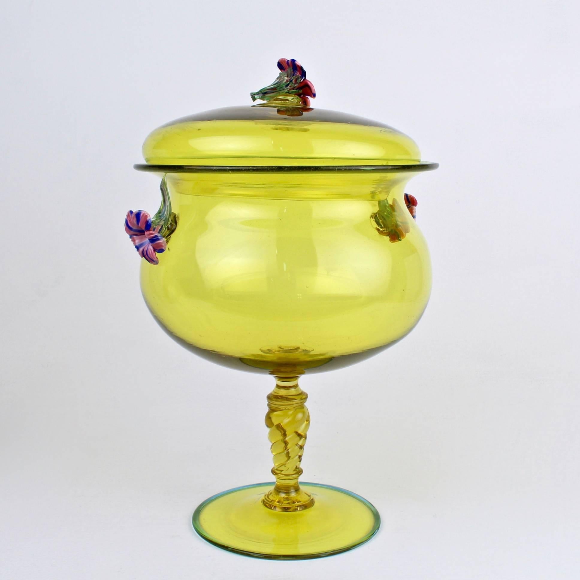 A fine, large, complex Venetian glass covered compote in canary yellow. 

The compote has a figural pink and blue applied finial and handles in the form of a trumpet shaped flowers. The lid rests on an urn from bowl that is supported by a twisted