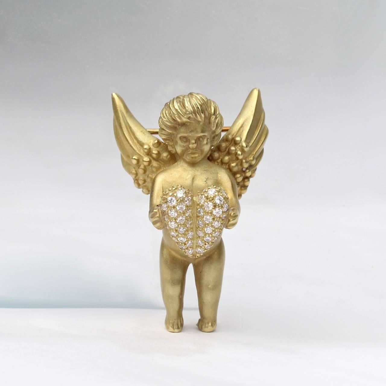 A charming variation on jewelry designer Barry Kieselstein-Cord's Angel or Cupid brooch. 

The cupid figure (or angel) stands with wings spread and outstretched hands holding a heart.

This variant has a matte gold finish and is set with over 80