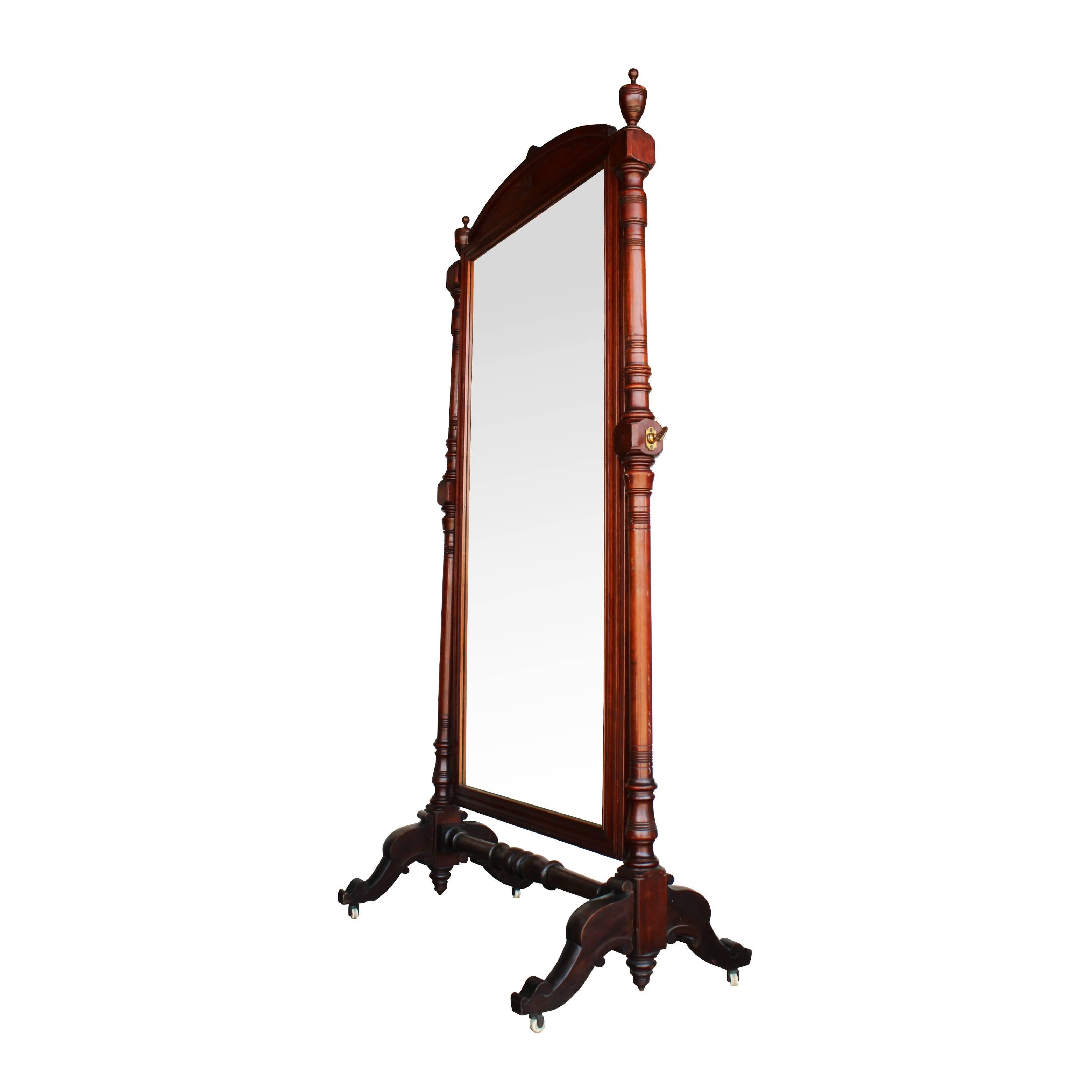 This elegant late 19th century mahogany cheval mirror features plenty of turned details on each supporting arm, brass hardware, and four porcelain casters for mobility. Urn finials and an arched header add flourished of additional detail to this