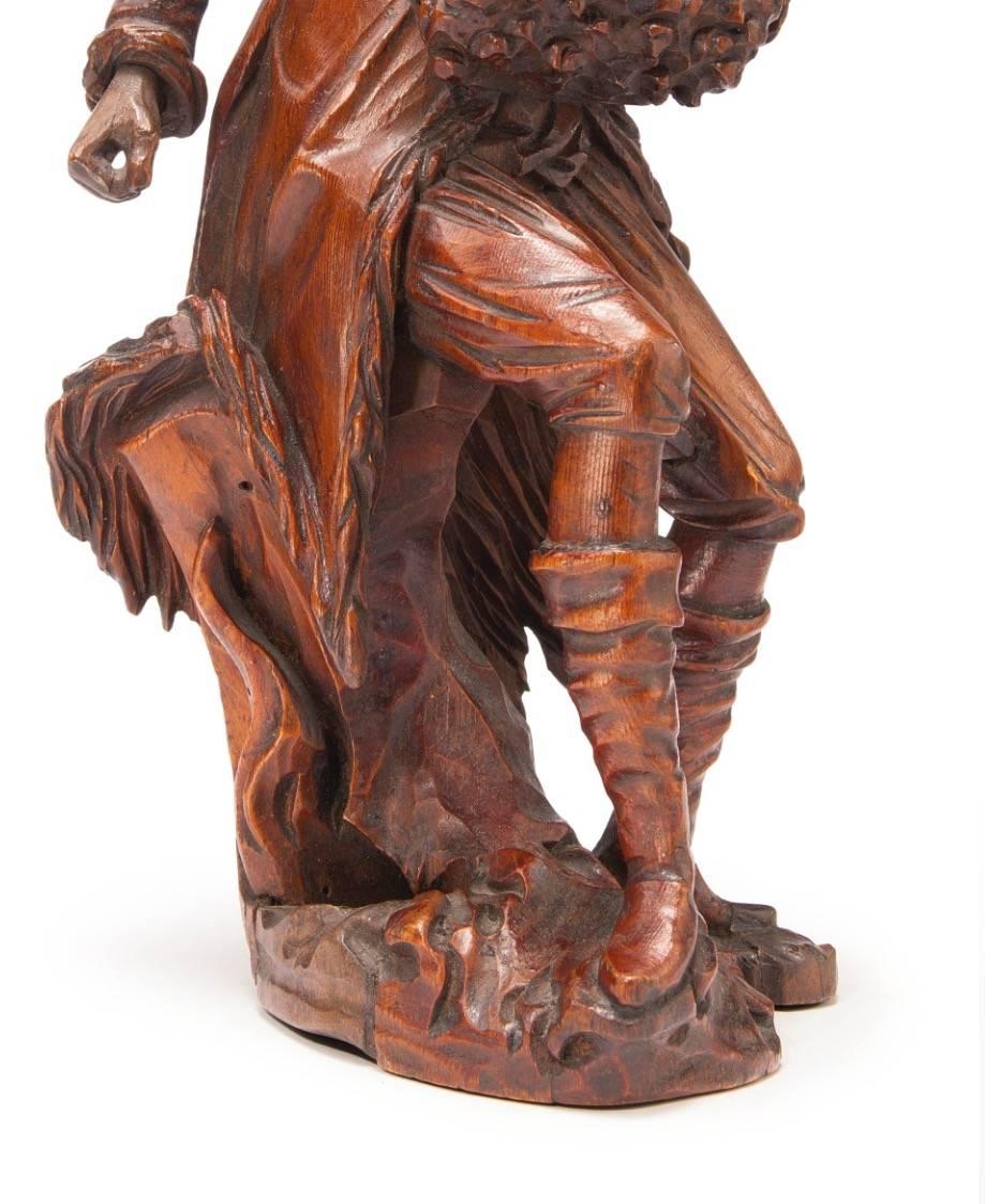 The carved figure with fur hat and muff, seated on a log and standing before a fire.