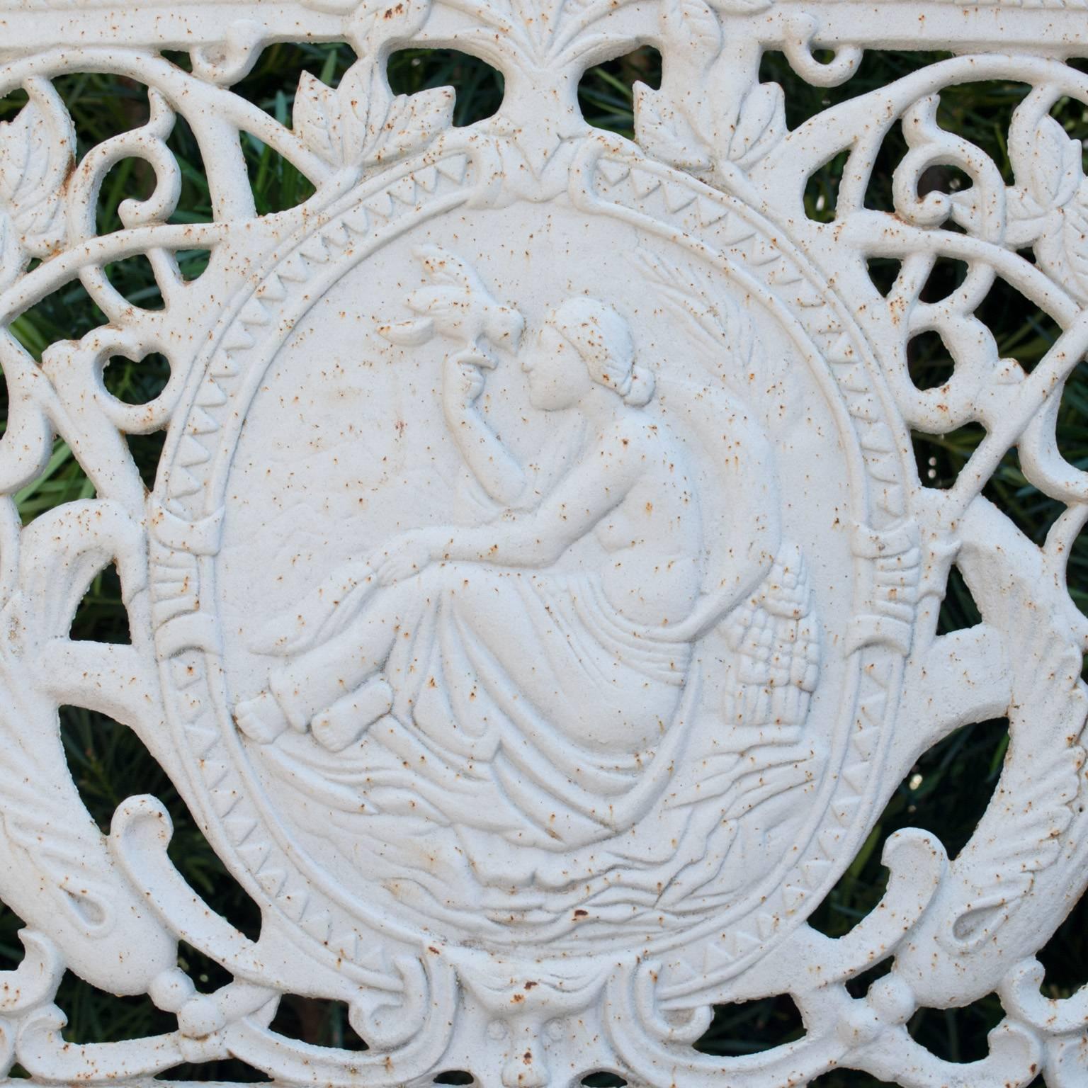 White cast iron bench with bowed legs, centered oval medallion depicting Venus and heavily ornate with foliage. Armrests are decorated and topped with sphere finials.