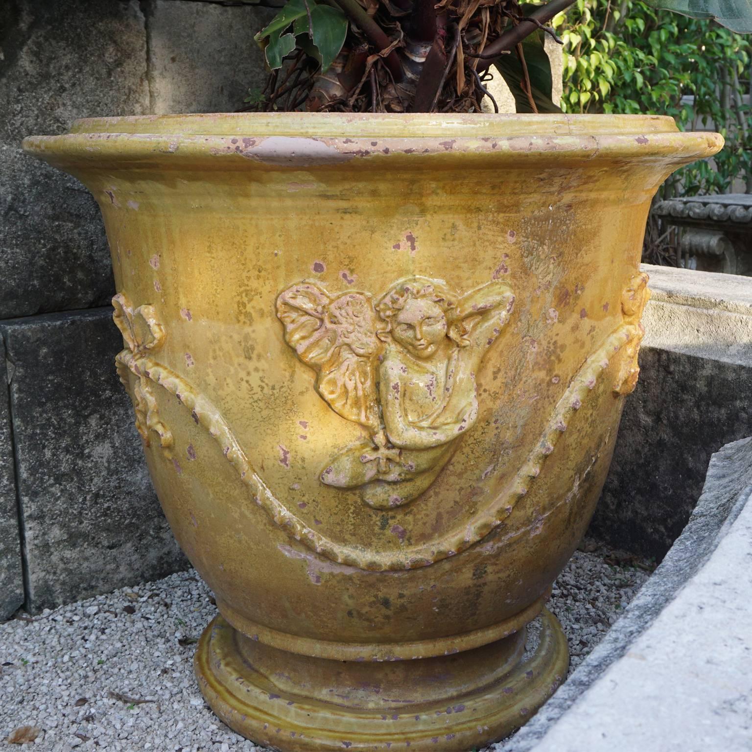 A vintage pair of Anduze urns has a more rustic and heavier appearance, originating from Provence in a glazed antiqued finish in a golden honey color. The planter is adorned with cherubs and ribbons. The finish is distressed and dimensions might