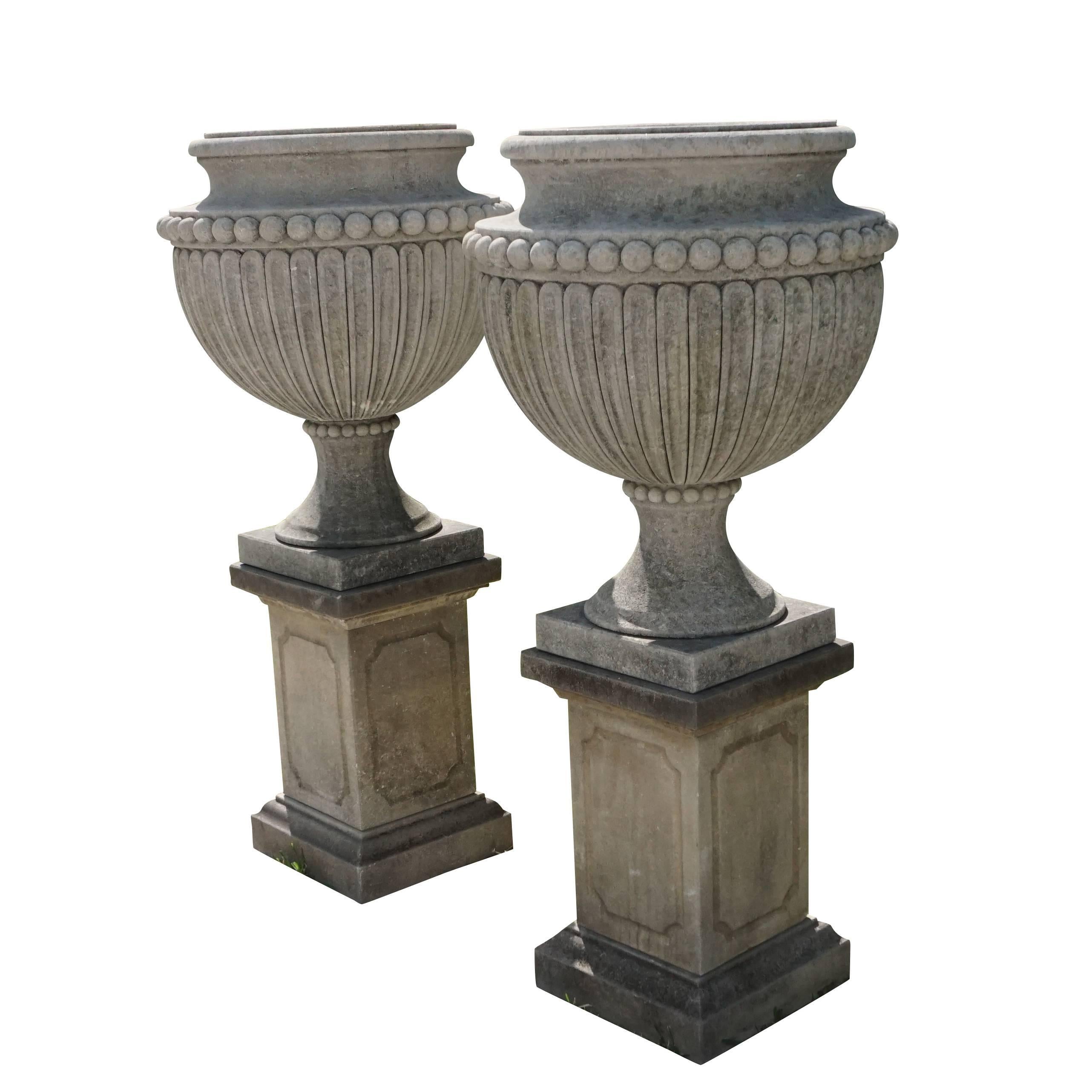 A pair of limestone urns “ Vases aux Gouttes “on original square pedestals with a fine bevel. The garden urns have a lobed ovoid body and are positioned on a circular foot with a square base. Wear consistent with age and use.

Urn: 32.5″ H

Base of
