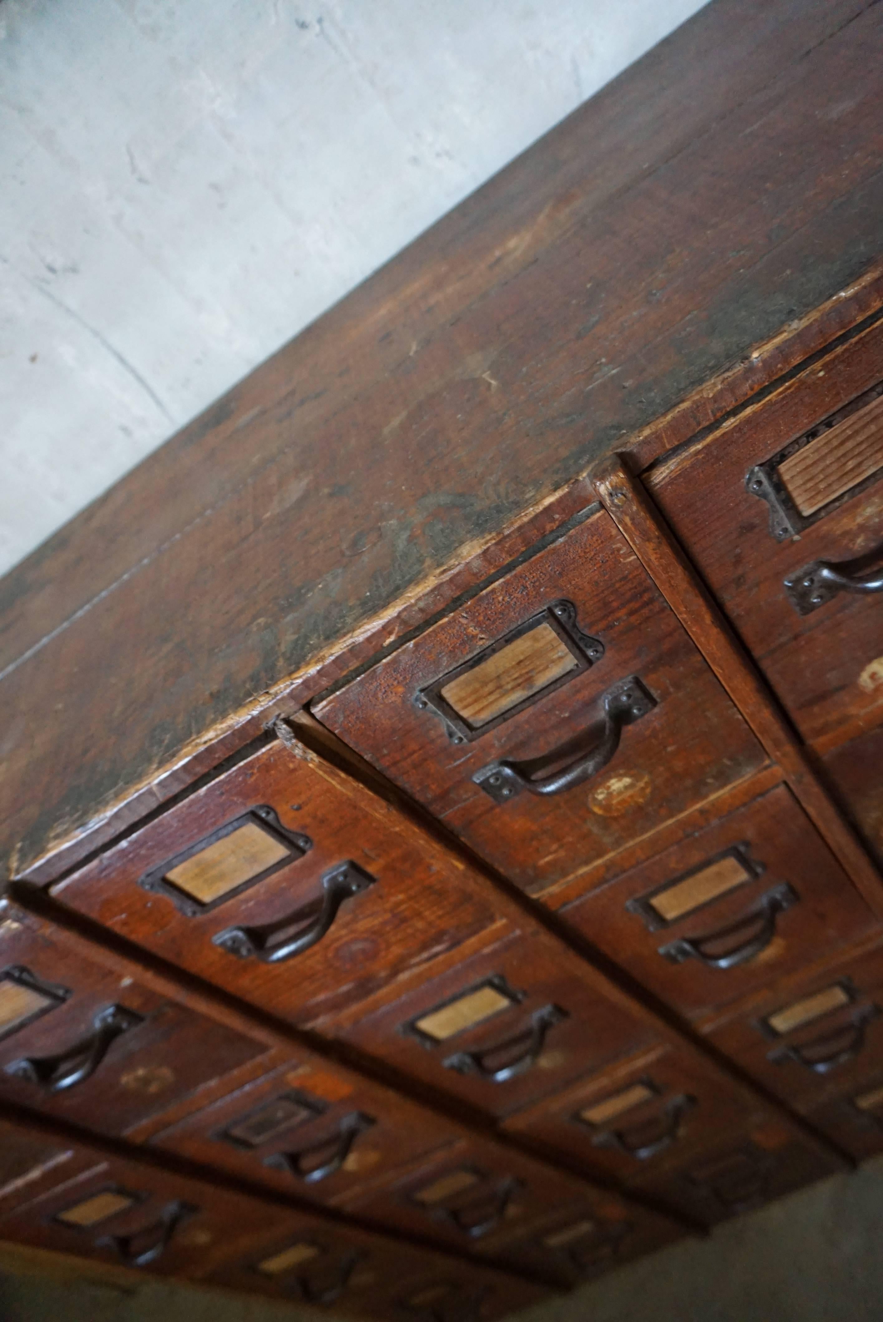 This apothecary bank of drawers was designed and made circa 1900. It is made from pine with iron handles. The cabinet has retained a rich patina with some natural distressing.