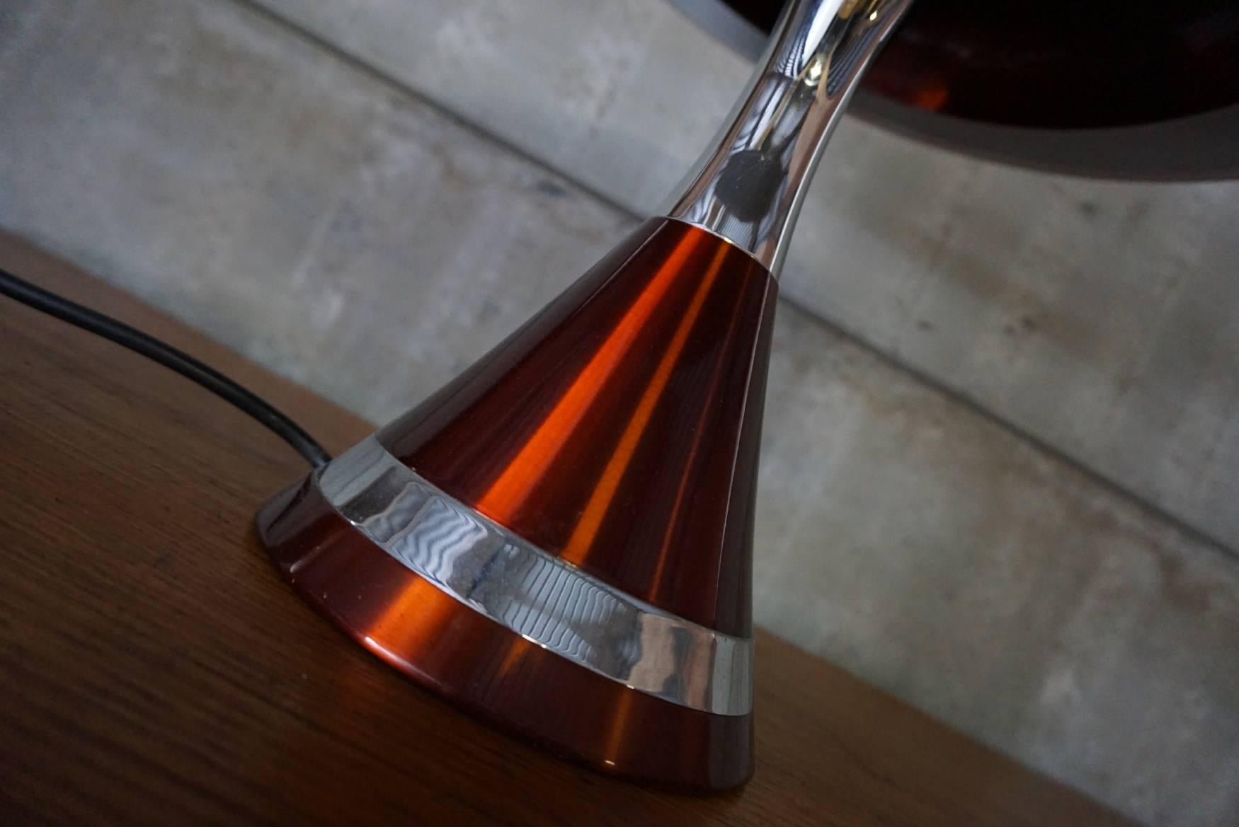 A space-age table lamp with a trumpet shaped base cast in bright orange or copper metallic. The outer shade is fitted on a white inner shade, trumpet shaped towards the bottom and open towards the top. The lamp features four bulb holders, one on top