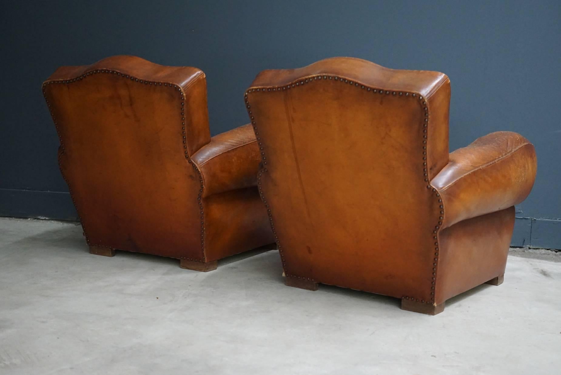 These club chairs were designed and produced in France during the 1940s. The chairs are made from cognac leather held together with metal pins and mounted on wooden legs. The chairs are in a vintage well used condition.
  