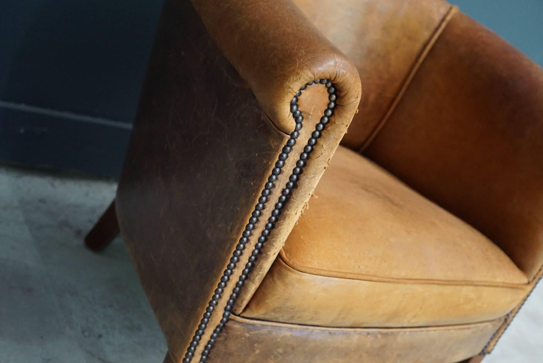 This club chair was designed and produced during the second half of the 20th century. The chair is made from cognac leather held together with metal pins and mounted on wooden legs. The lounge chair is in a vintage well used condition.