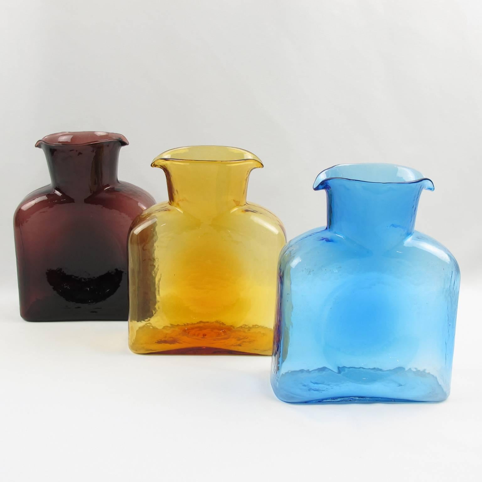Lovely vintage set of three pitchers, jugs, decanters by Blenko Glass Manufacturer. Square geometric shape with double spouted design. Assorted colors of purple amethyst, orange amber and blue sky. The amber color has original etched Blenko marking
