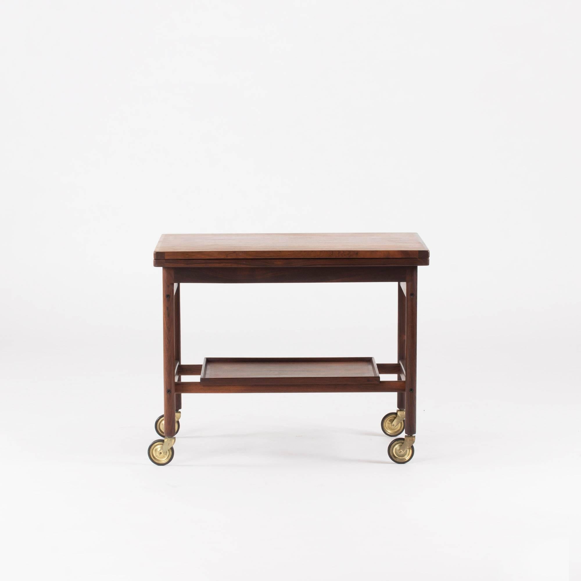 Elegant rosewood bar trolley by Kurt Østervig with a table top that folds up to become double length. The inside is covered with black formica. Removable tray underneath the top with a beautiful pattern in the rosewood. Brass wheels.

Length when