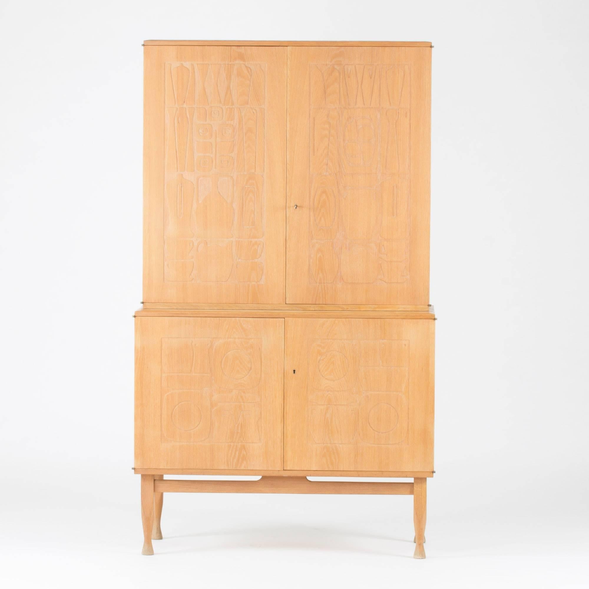"Krus" ("Vessel") cabinet by Yngve Ekström, in a rare edition in washed oak and with sculpted legs. The front doors display a modernistic relief of everyday kitchen objects mixed with flowers and decorative objects. An arty,