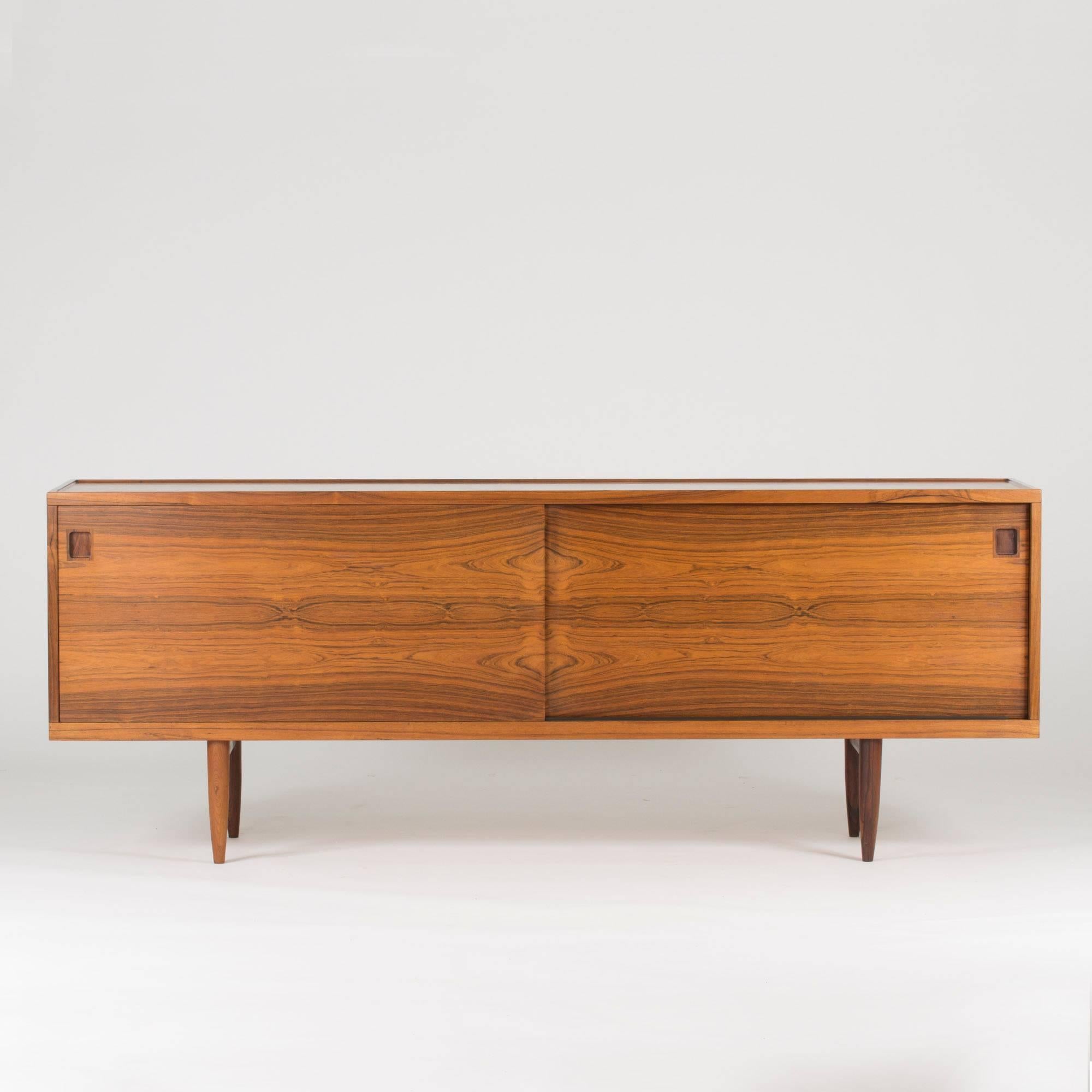 Exquisite rosewood sideboard by Niels O. Møller in a smooth design where the beautiful wood grain falls into focus on the two sliding doors. The top is carved in a soft curve around the edge, giving this piece an extra dimension that at first can go