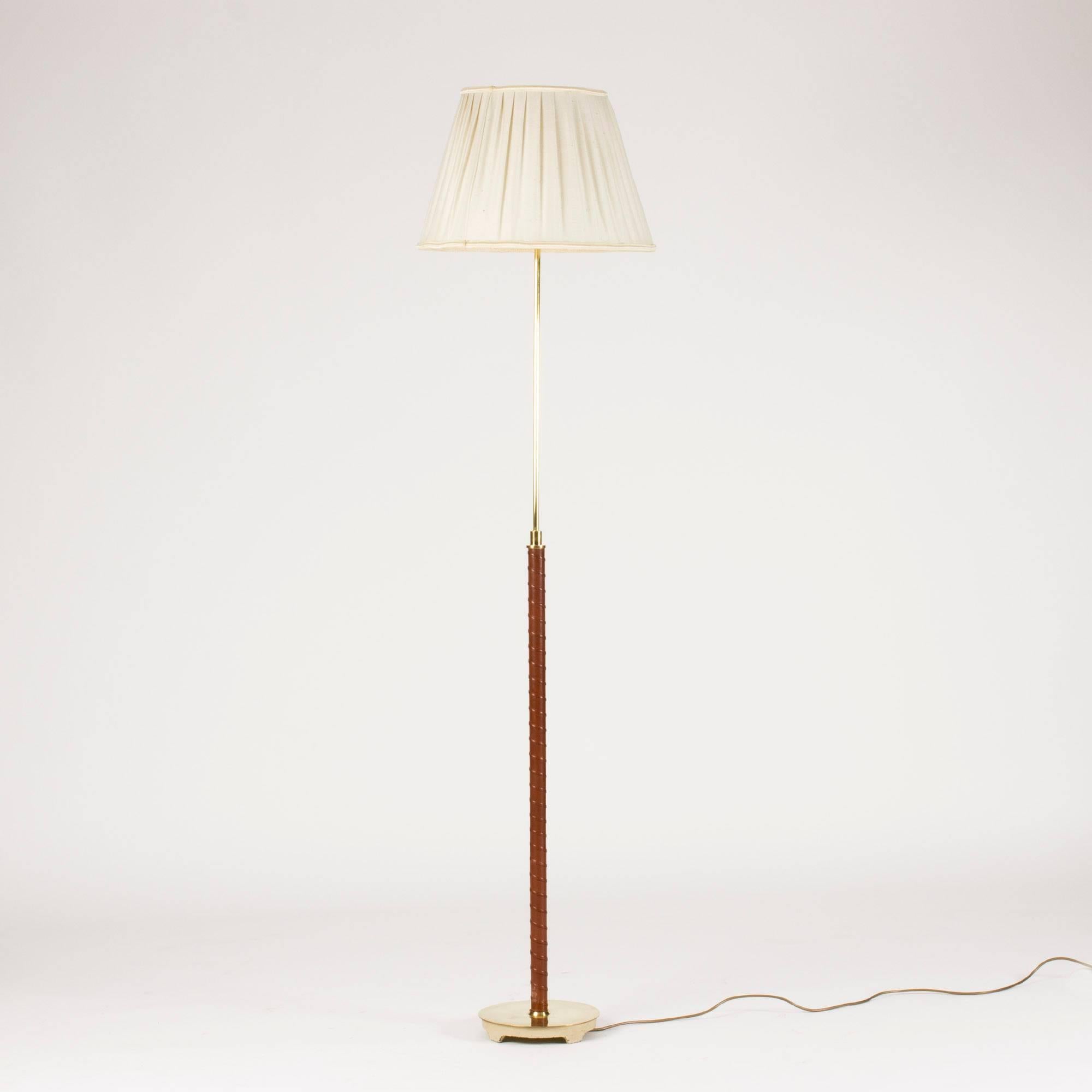 Elegant floor lamp from NK, with a brass base with sculpted feet and handle wound with brown leather. The height of the lamp is adjustable vertically. Original textile shade in good condition, somewhat yellowed.