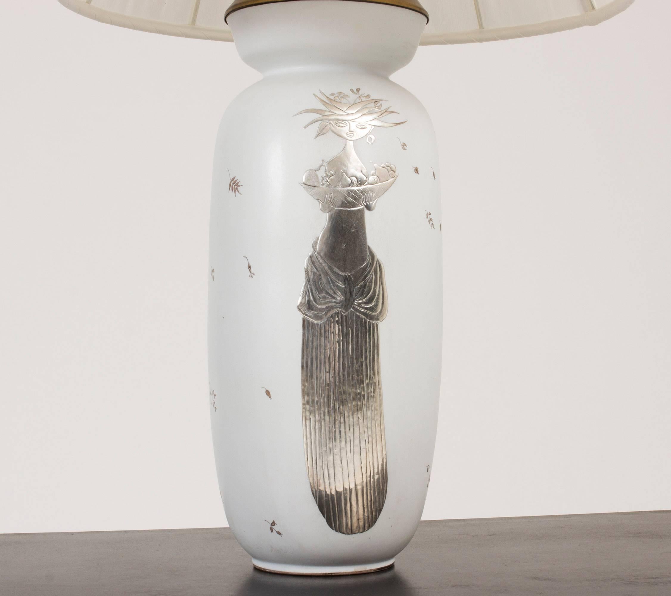 Beautiful, tall “Grazia” table lamp by Stig Lindberg made from Carrara stoneware. Exquisite silver decor of a woman holding a basket full of fruits. White textile shade reupholstered on the original lamp shade frame.

Measure: The height of the