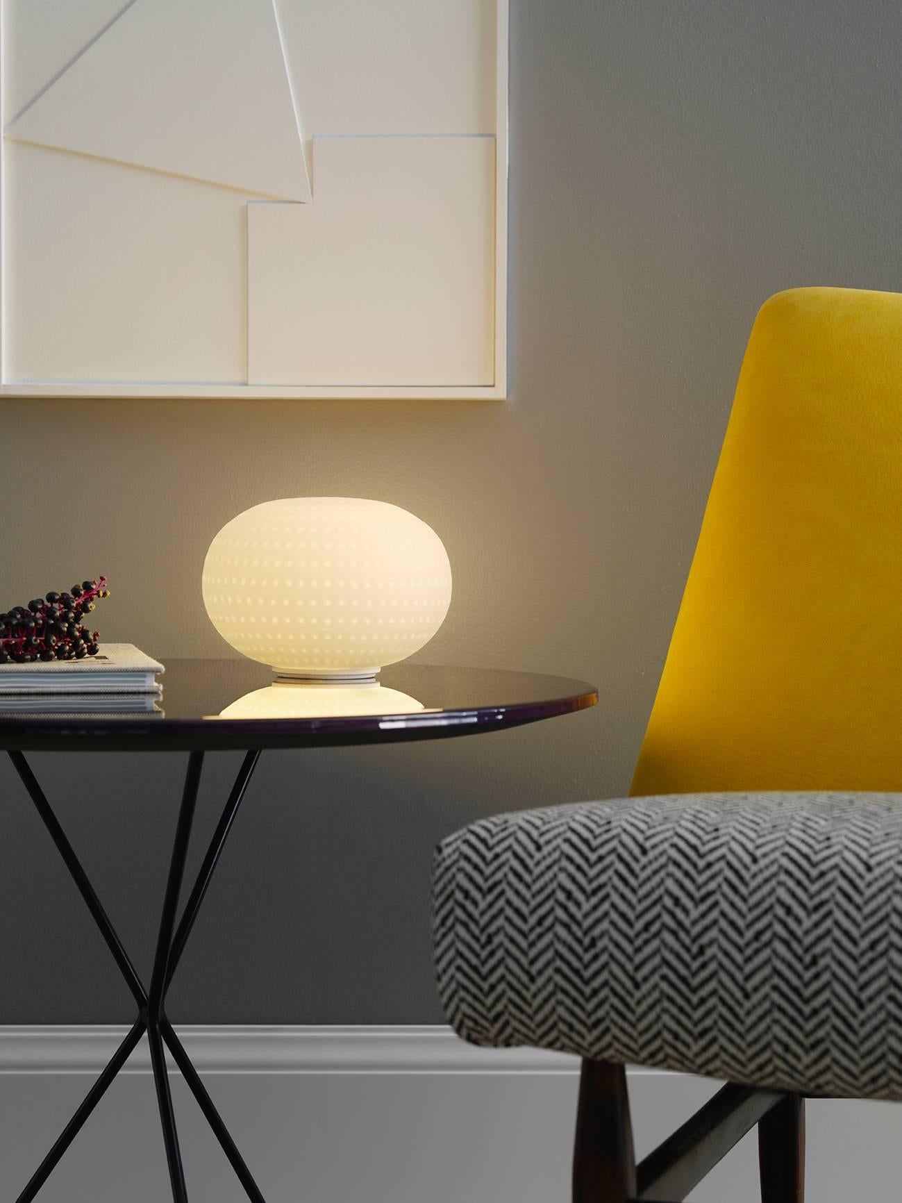 Bianca small table lamp without stem by Matti Klenell from Fontana Arte.

Its beautifully balanced shape is enriched and made unique by an orderly series of delicate, light grooves that, on the white volume of the glass, look like tiny prints on