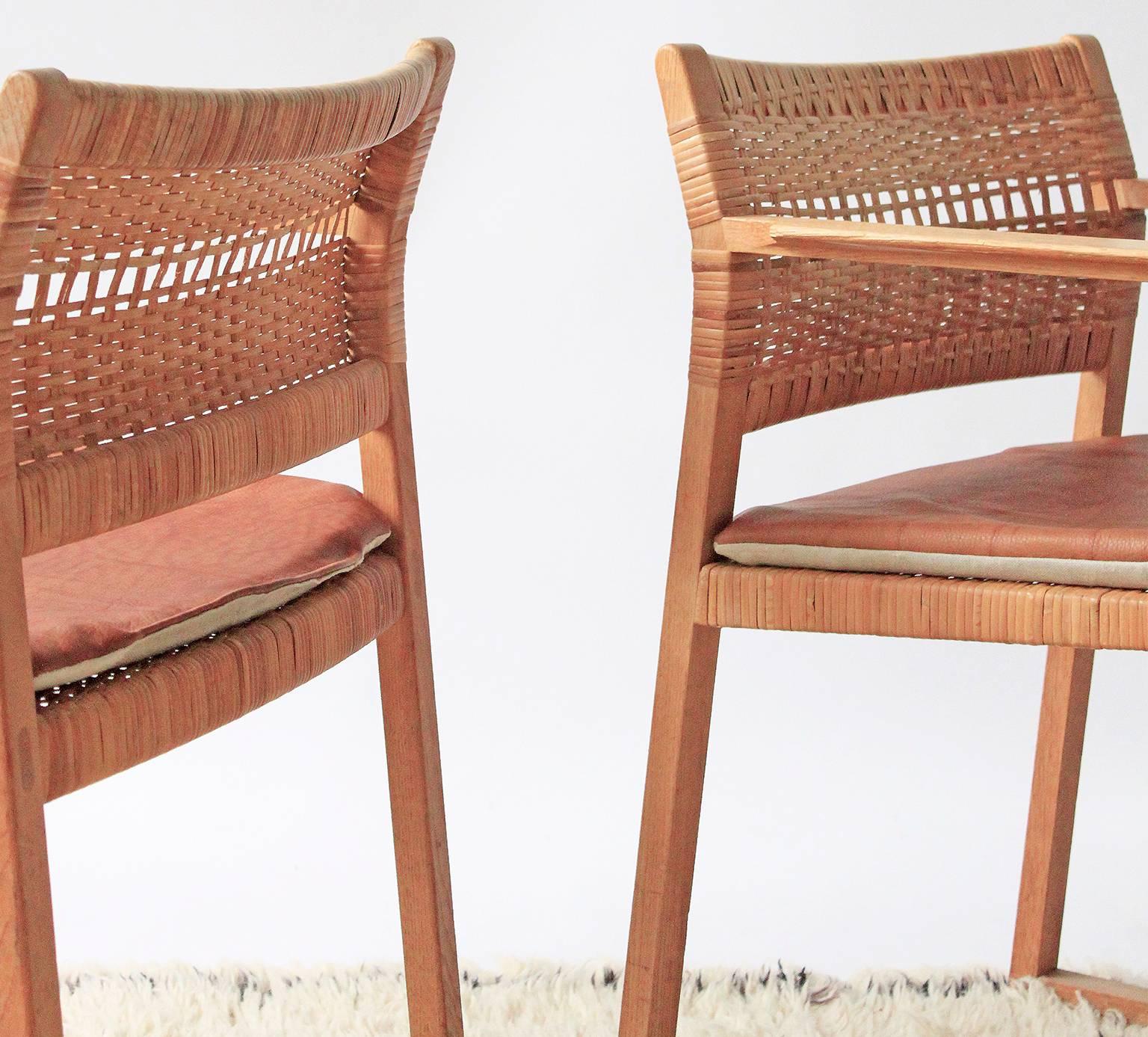 A pair of model BM65 dining chairs. This is the variant with arms, an oak frame and woven cane seat and back. Chairs include new reversible cushions made with natural leather and Belgian linen.  Some cane damage and some strands of cane repaired of