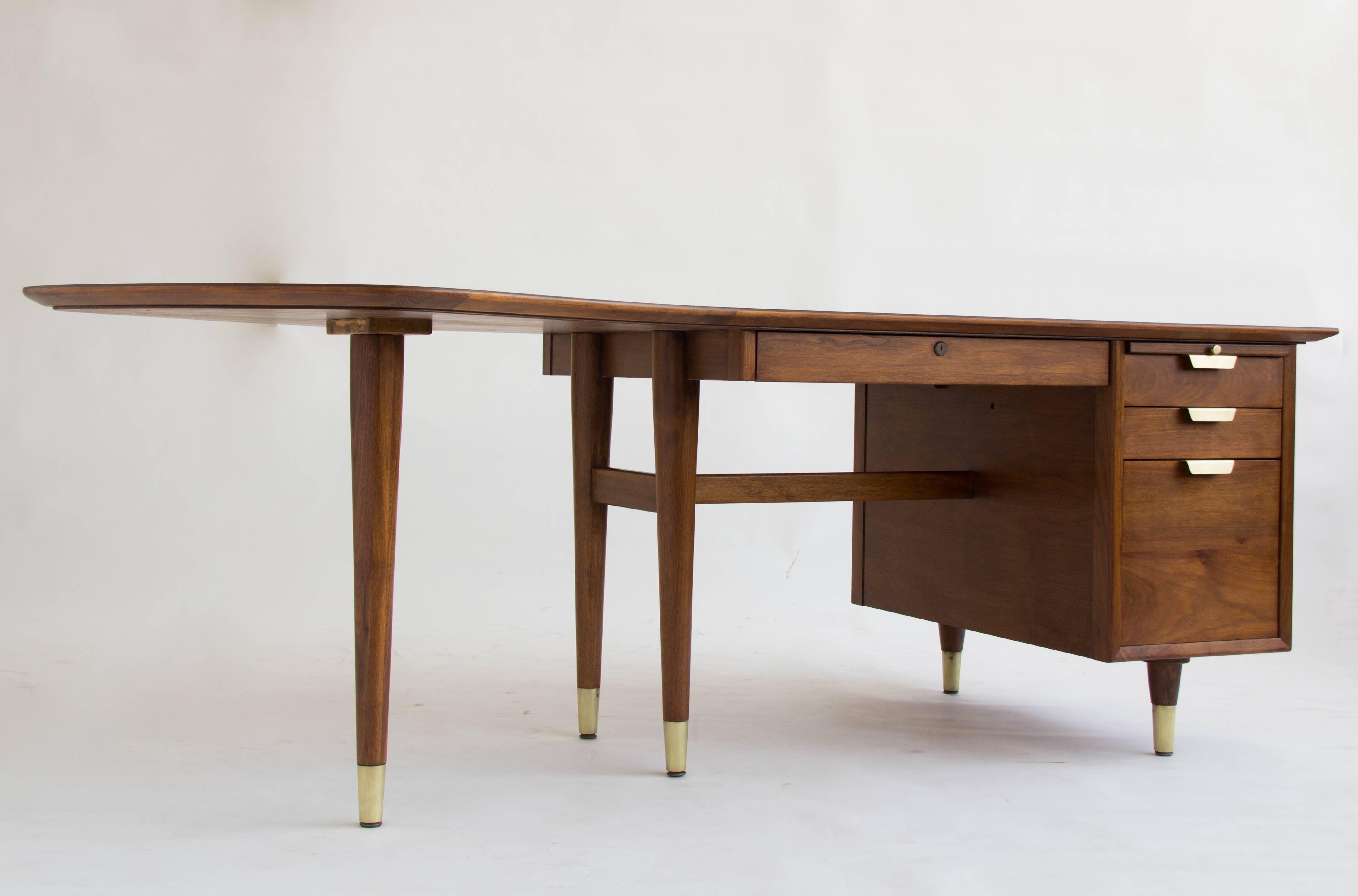 This walnut executive desk features a unique boomerang-shaped left return. The table surface is walnut and is edged with a band of contrasting grain wood. A shared mechanism locks all three right-hand drawers when the bolt on the center drawer is